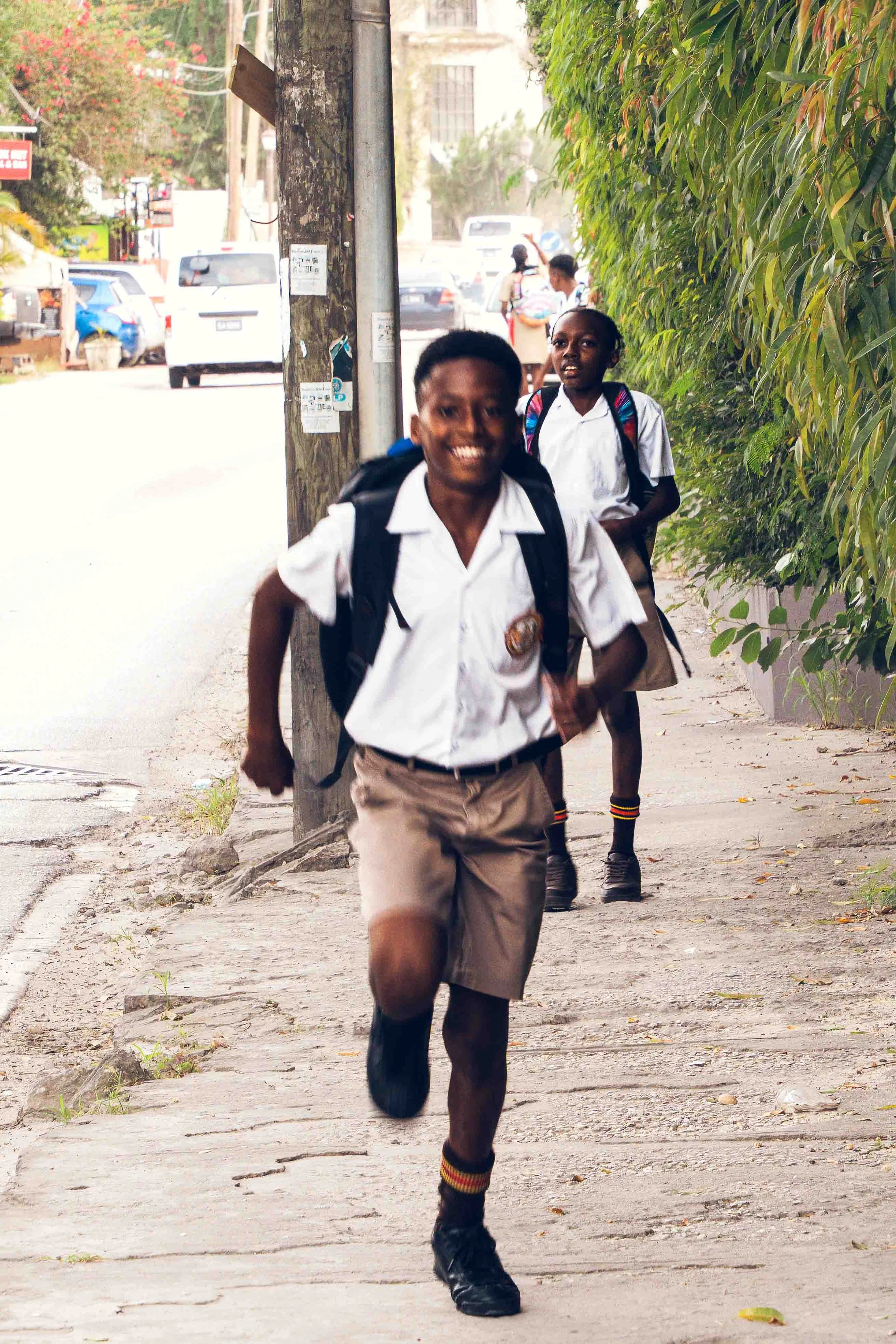 Photographie documentaire montrant des élèves en uniforme scolaire marchant et courant sur un trottoir après l’école. Les enfants portent des sacs à dos et se déplacent le long d’une rue bordée de végétation dans un environnement tropical. L’image ca