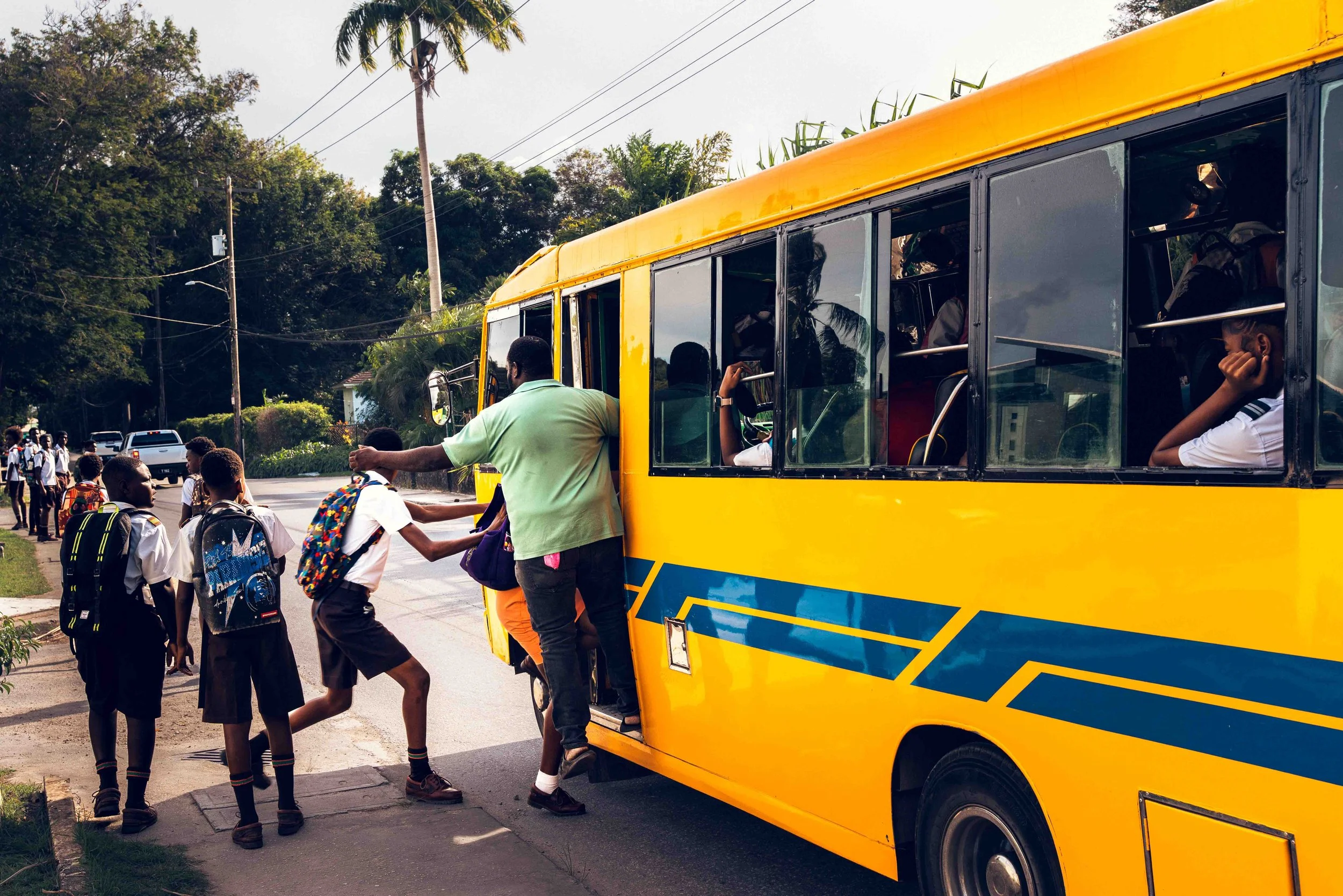 Photographie documentaire montrant des élèves montant dans un bus scolaire jaune dans un environnement tropical. La scène capture un moment du quotidien à la sortie de l’école, avec des enfants en uniforme, des sacs à dos colorés et l’animation de la