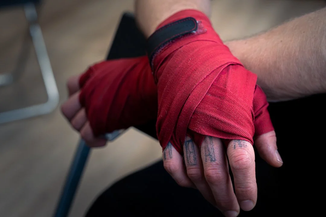 Photographie de reportage montrant les mains d’un boxeur enveloppées dans des bandes rouges avant un combat ou un entraînement. Le cadrage serré met en évidence les détails des protections, la texture des bandages et les tatouages visibles sur les do