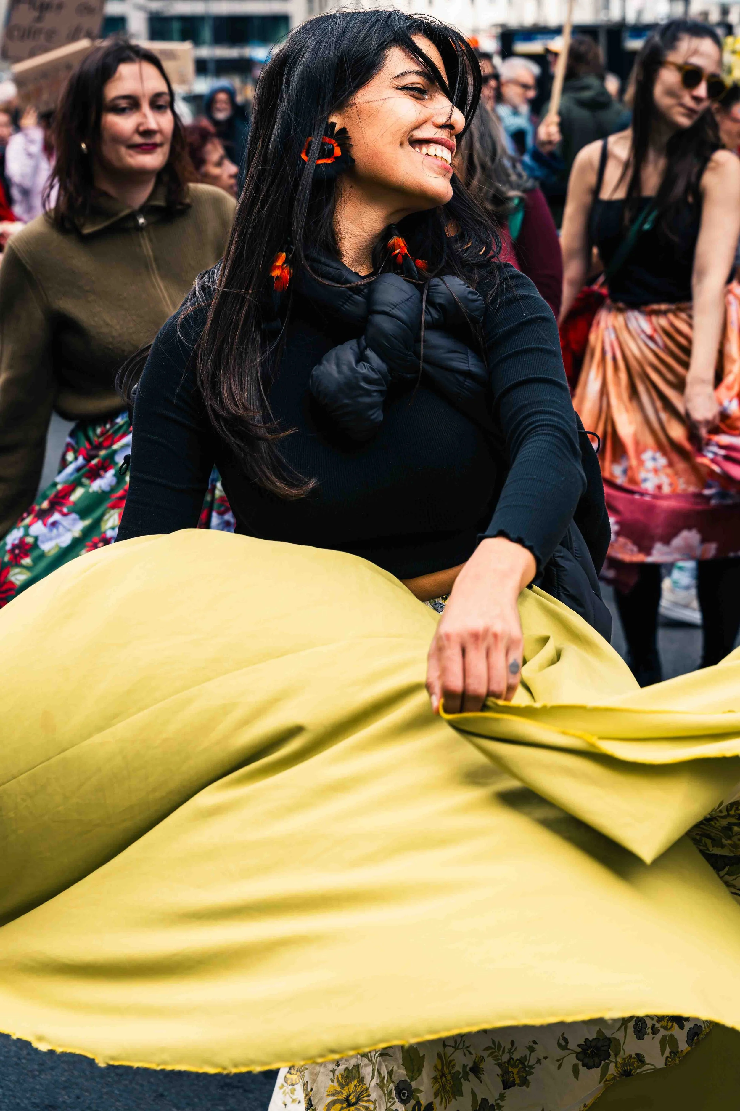 Photographie de reportage réalisée dans l’espace urbain lors d’une manifestation à Bruxelles. Une femme danse au cœur du cortège, entraînant un large tissu jaune dans son mouvement tandis que d’autres participantes défilent derrière elle. L’image cap