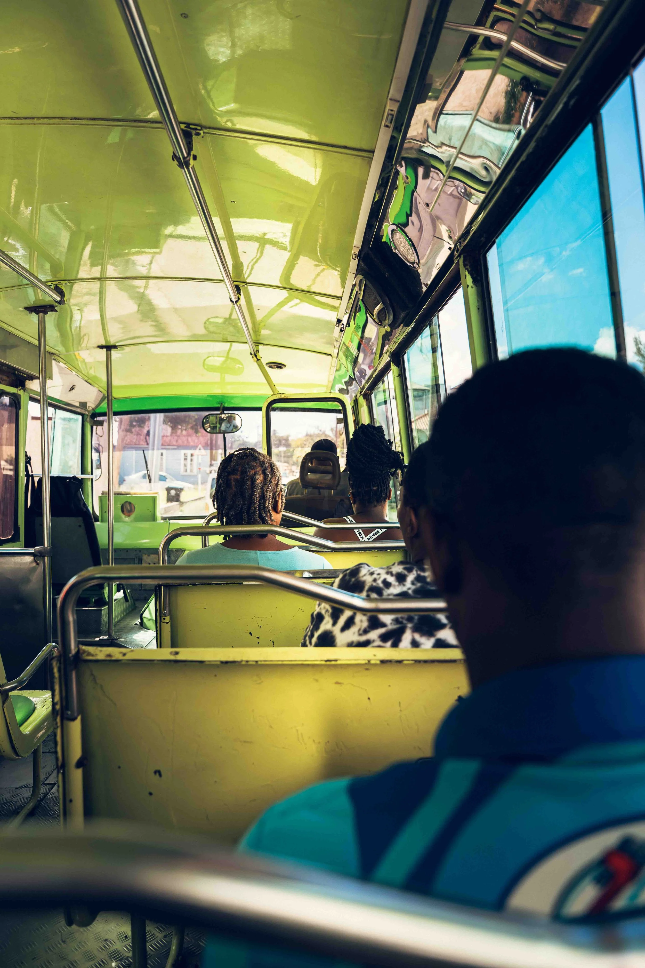 Photographie documentaire prise à l’intérieur d’un bus en circulation, montrant des passagers assis et observés depuis l’arrière du véhicule. L’image met en valeur la perspective des sièges, la lumière naturelle entrant par les fenêtres et les couleu