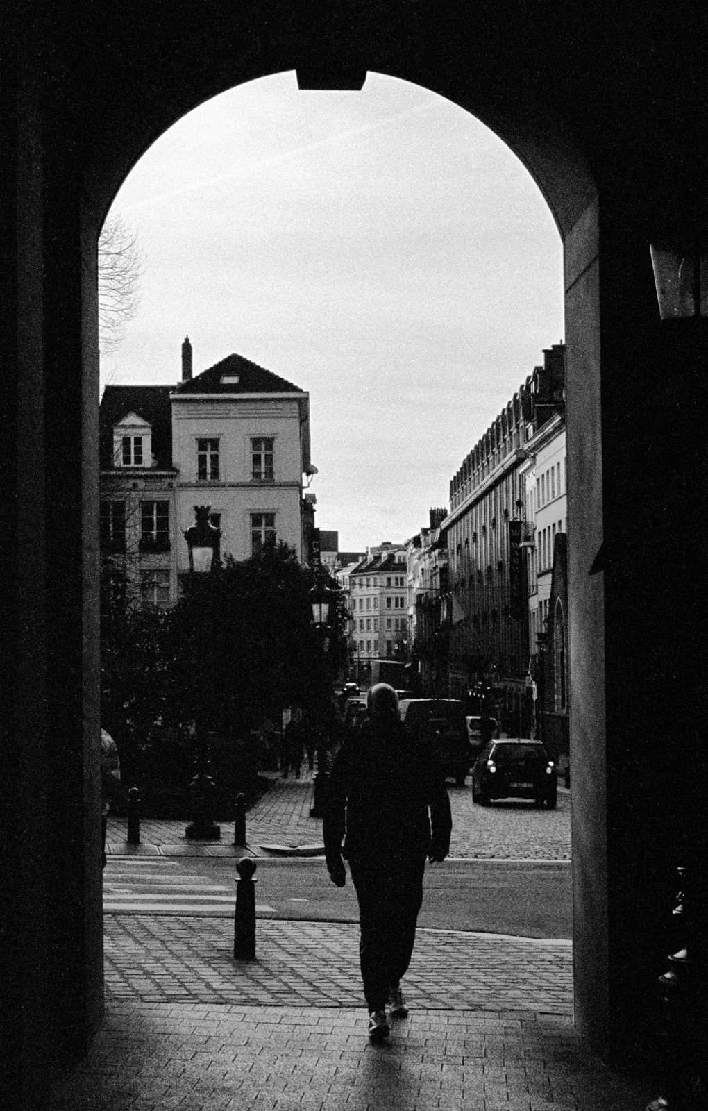Photographie en noir et blanc d'une silhouette traversant un passage voûté ouvrant sur une rue de Bruxelles, mettant en valeur le cadrage architectural.
