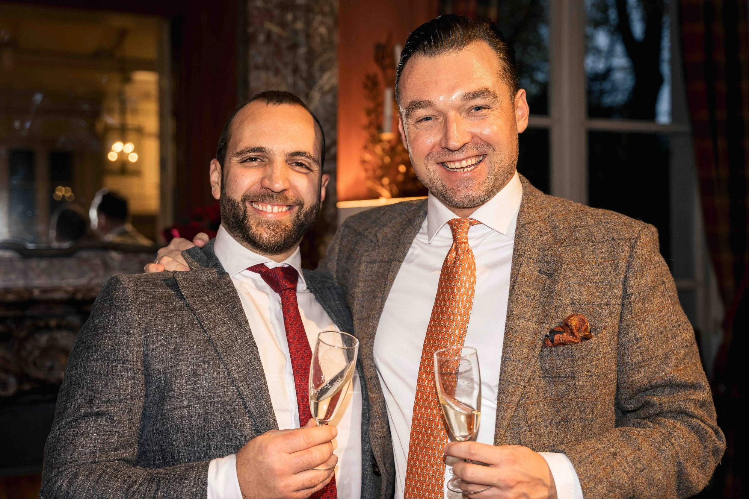 Photographie d’événement montrant deux hommes élégamment habillés en costume levant des coupes de champagne lors d’une réception. Les deux invités sourient face à l’appareil dans un intérieur raffiné éclairé par une lumière chaleureuse, évoquant une 