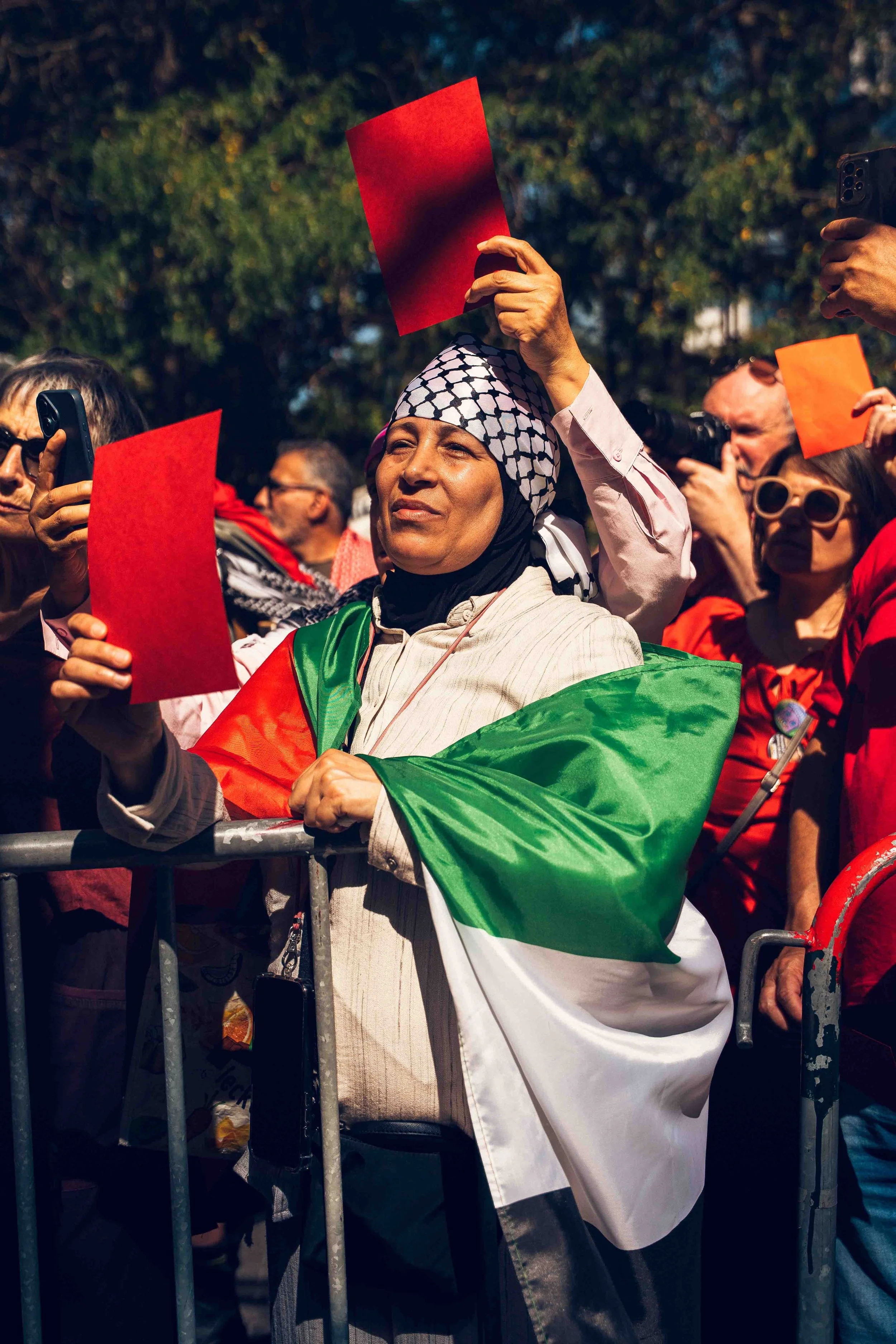 Photographie de reportage capturant une manifestante tenant un carton rouge lors d’un rassemblement pro-palestinien. Drapée dans un drapeau palestinien et entourée d’autres participants, elle se tient derrière une barrière de sécurité au cœur d’une m