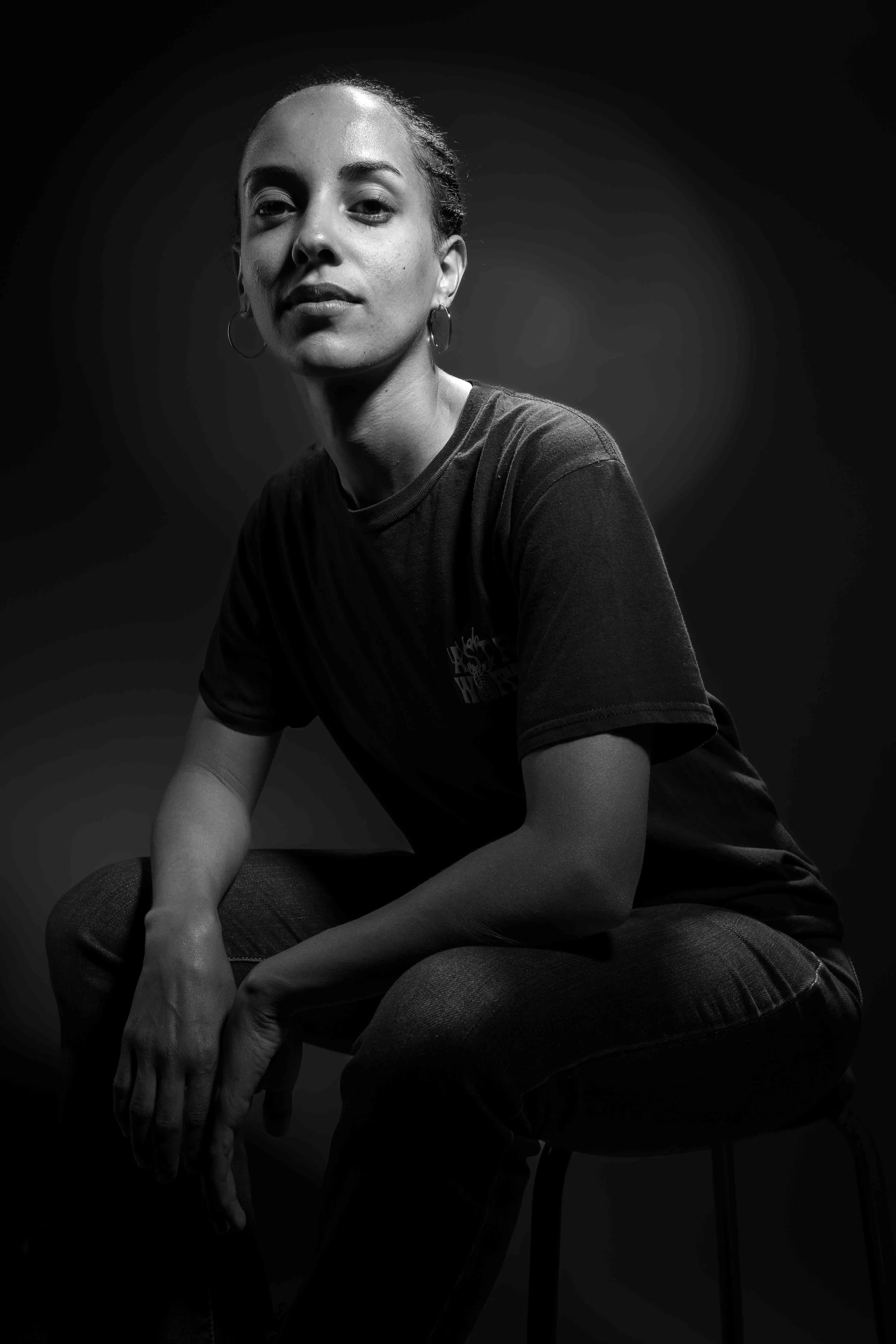 Portrait en noir et blanc réalisé en studio à Bruxelles. Une jeune femme assise sur un tabouret regarde l’objectif avec calme et assurance. La lumière latérale sculpte le visage et le corps, créant un contraste marqué entre ombre et lumière. L’image 