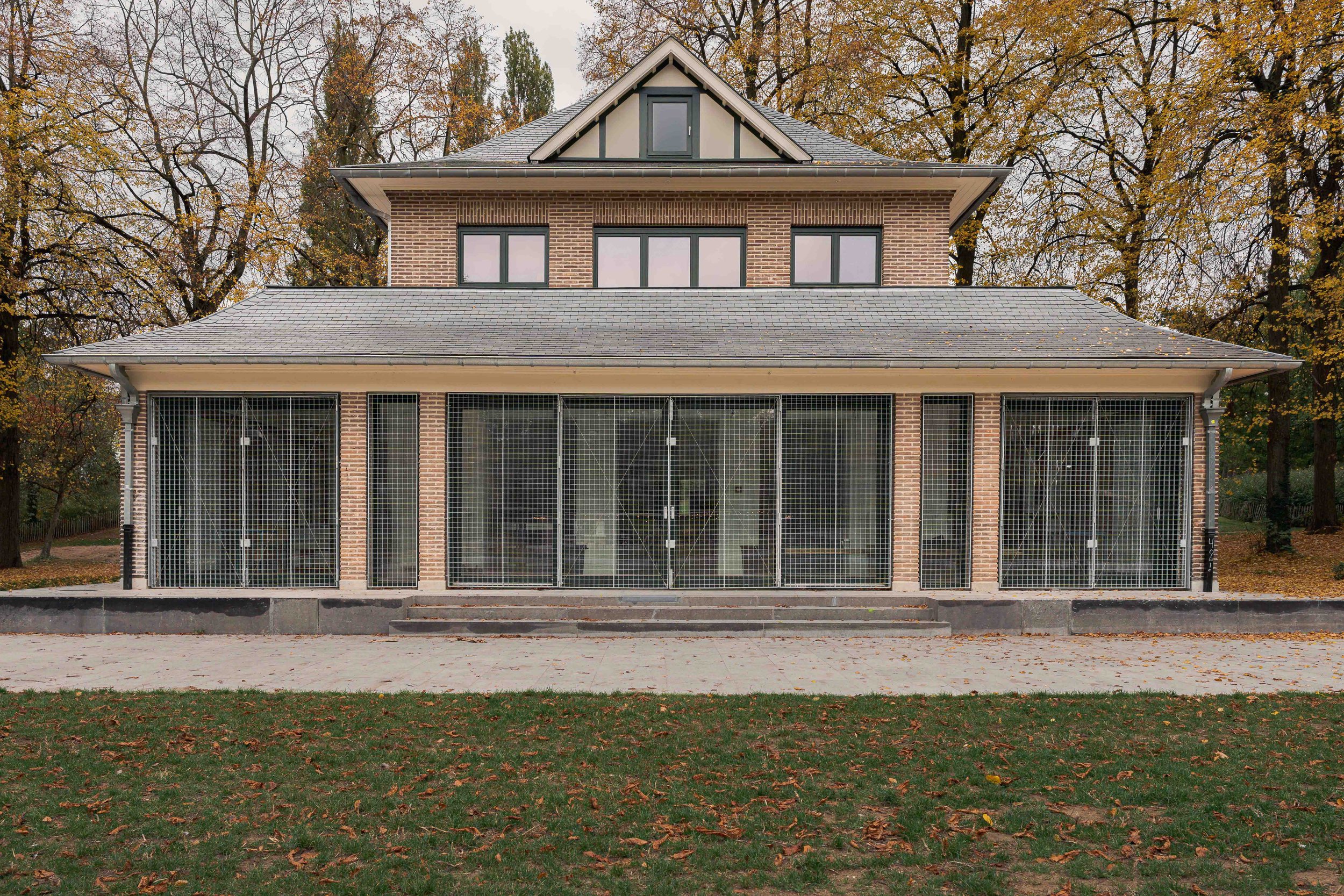 Photographie d’architecture montrant un pavillon symétrique en briques situé dans un parc arboré en automne. La façade régulière composée de grandes baies vitrées protégées par des grilles métalliques et le toit à double pente créent une composition 