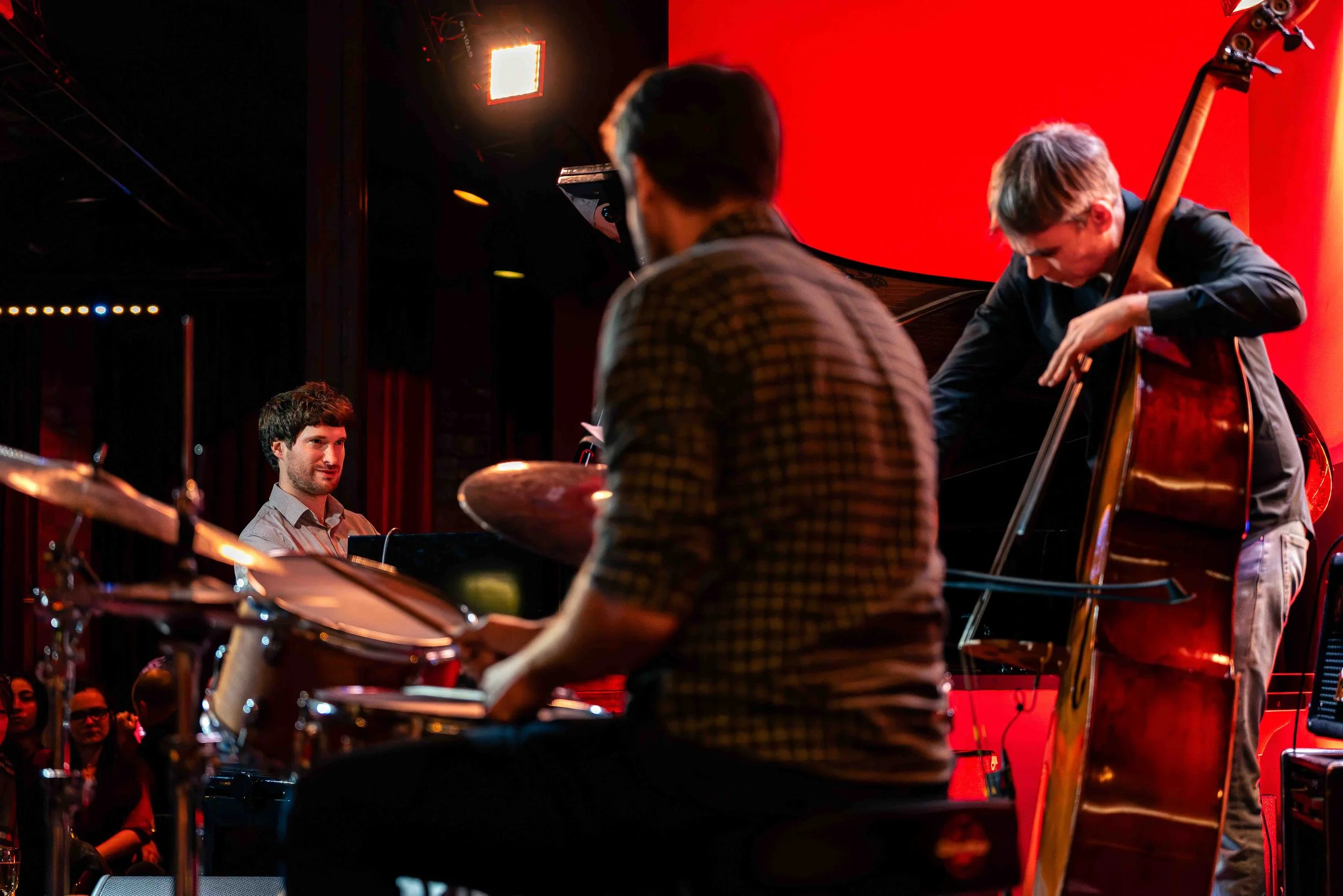 Photographie de reportage capturant un trio de jazz en concert sur scène. Un batteur, un pianiste et un contrebassiste jouent ensemble sous un éclairage de scène rouge, dans l’atmosphère intime d’un club ou d’une salle de concert. L’image saisit l’in