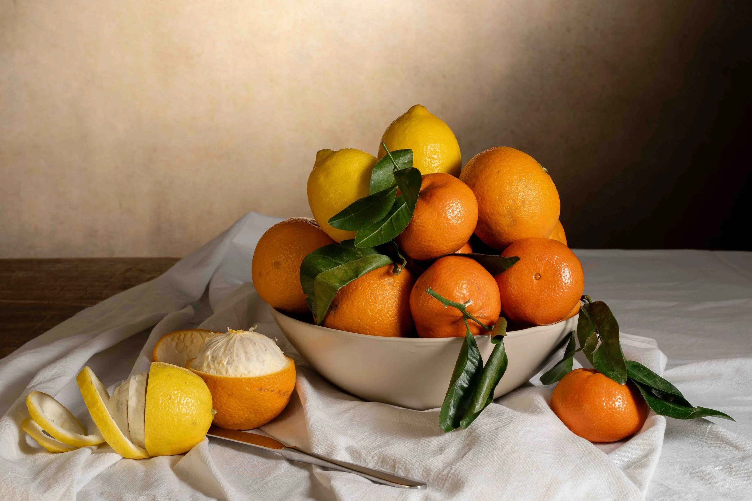 Photographie de nature morte montrant un bol rempli d’oranges et de citrons disposés sur une table recouverte d’un tissu clair. Les fruits frais avec leurs feuilles vertes créent une composition classique inspirée de la peinture, avec une lumière dou