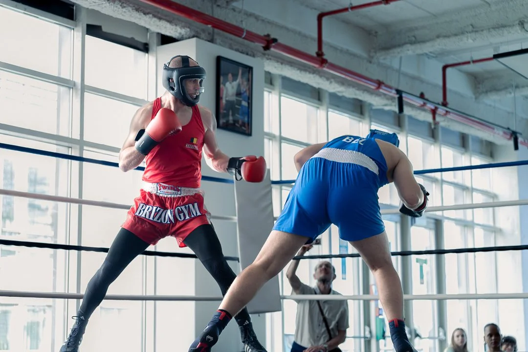 Photographie de reportage capturant un combat de boxe amateur dans une salle d’entraînement. Deux boxeurs équipés de gants et de protections s’affrontent sur le ring sous la lumière naturelle entrant par de grandes fenêtres. L’image documente l’inten
