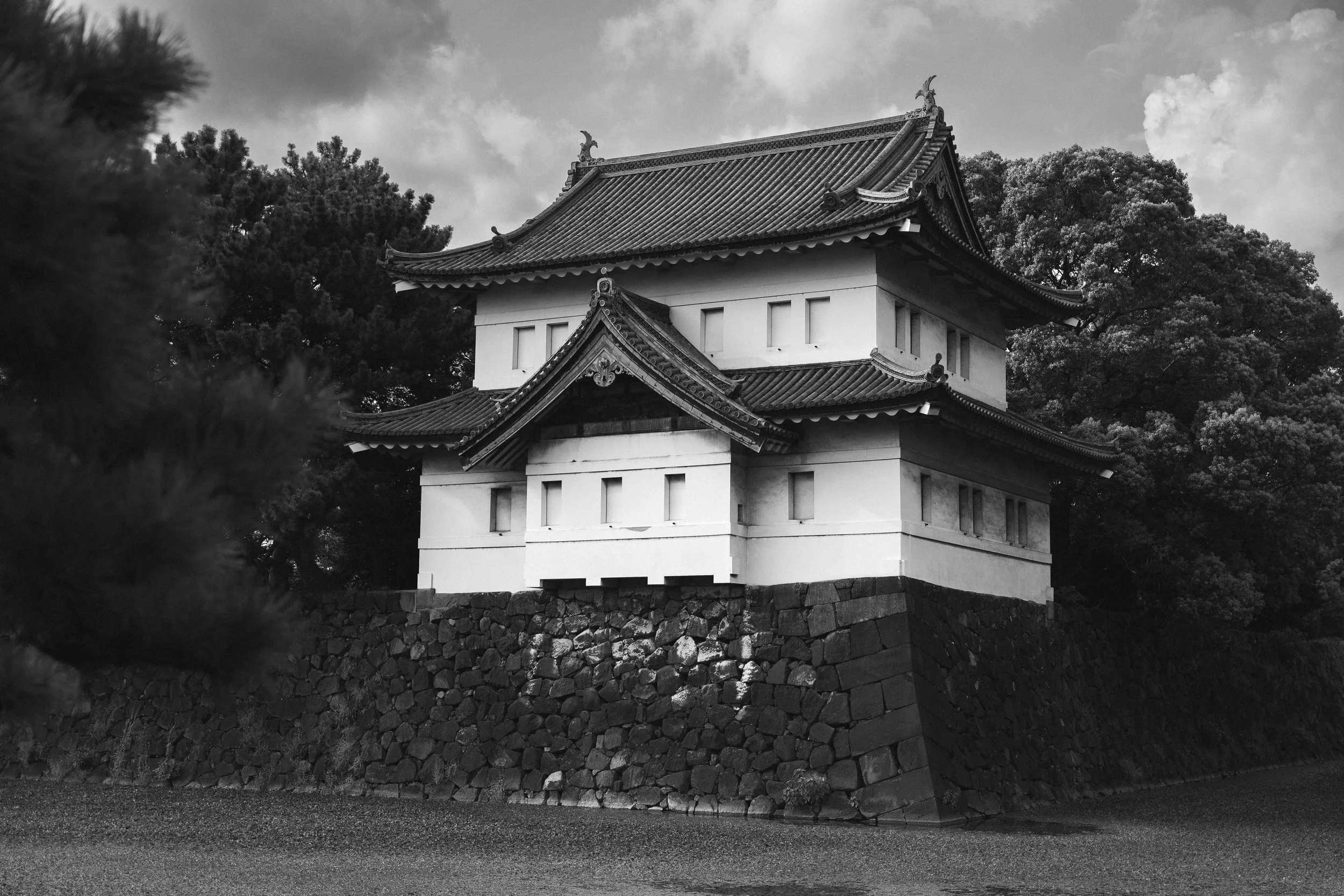 Photographie en noir et blanc d’un bâtiment traditionnel japonais ressemblant à une tour de garde ou à un pavillon de château, construit sur une base de pierres massives. L’architecture aux toits courbés typiques du Japon se détache sur un ciel nuage