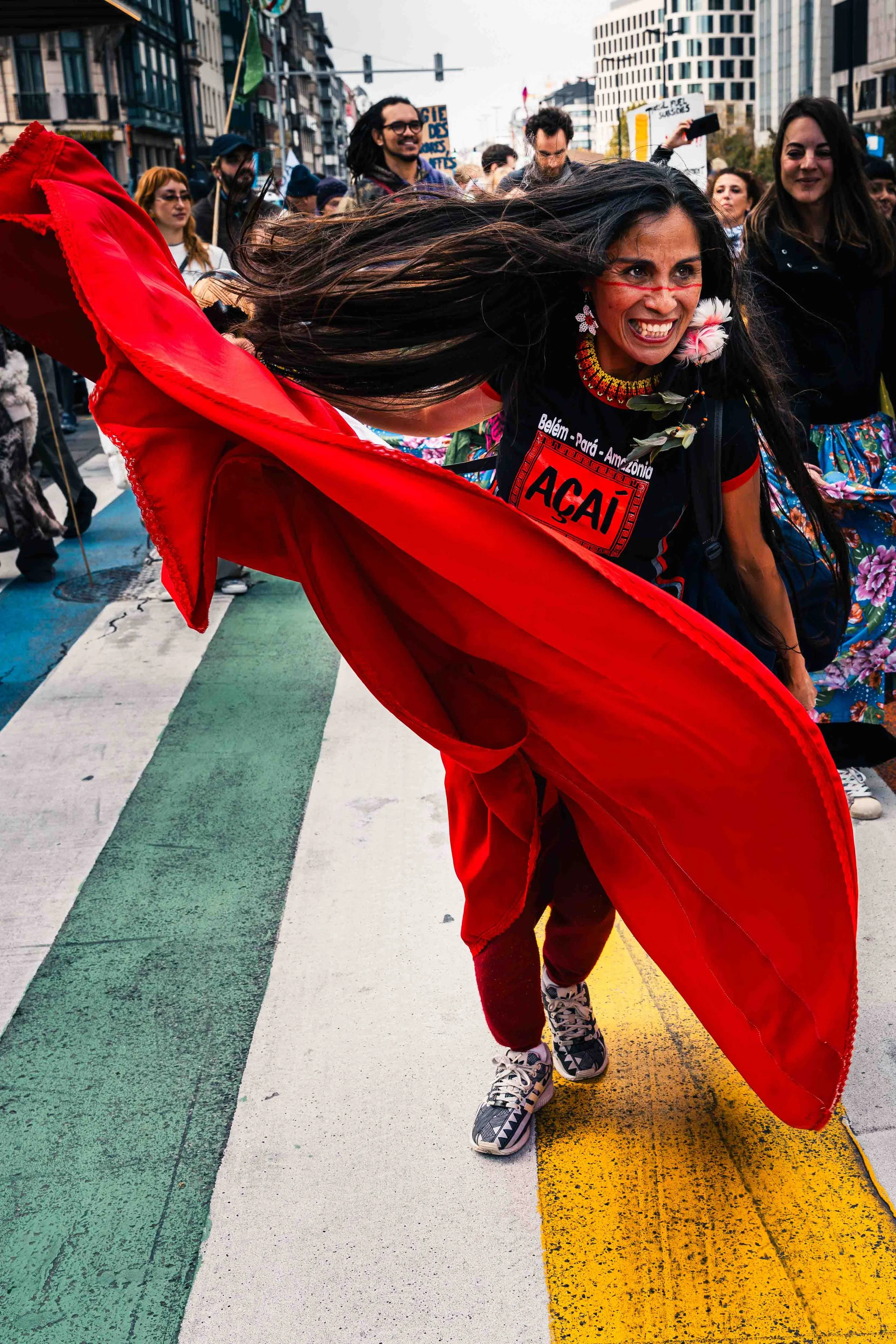 Photographie de reportage capturant une manifestation dans l’espace urbain à Bruxelles. Au premier plan, une femme vêtue de rouge avance avec énergie, un tissu flottant dans le mouvement, tandis qu’un groupe de manifestants la suit dans la rue. L’ima