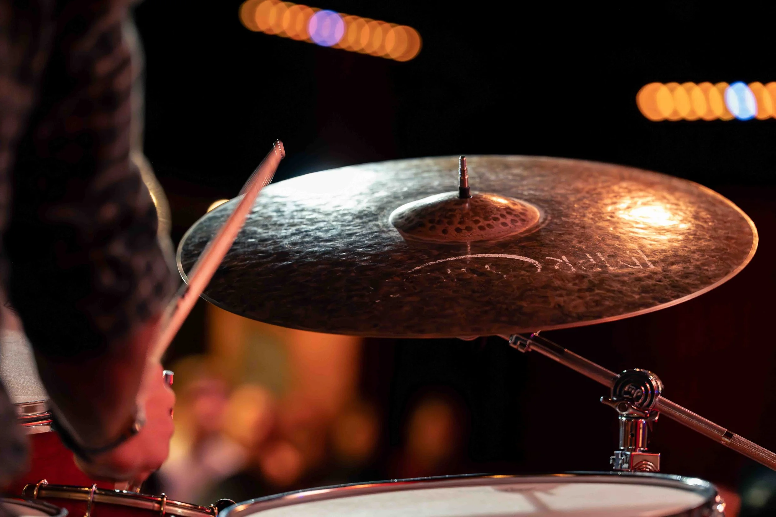 Photographie de reportage montrant un gros plan sur une cymbale de batterie frappée par une baguette lors d’un concert de jazz. La lumière chaude de la scène se reflète sur le métal martelé de l’instrument tandis que l’arrière-plan reste flou, évoqua