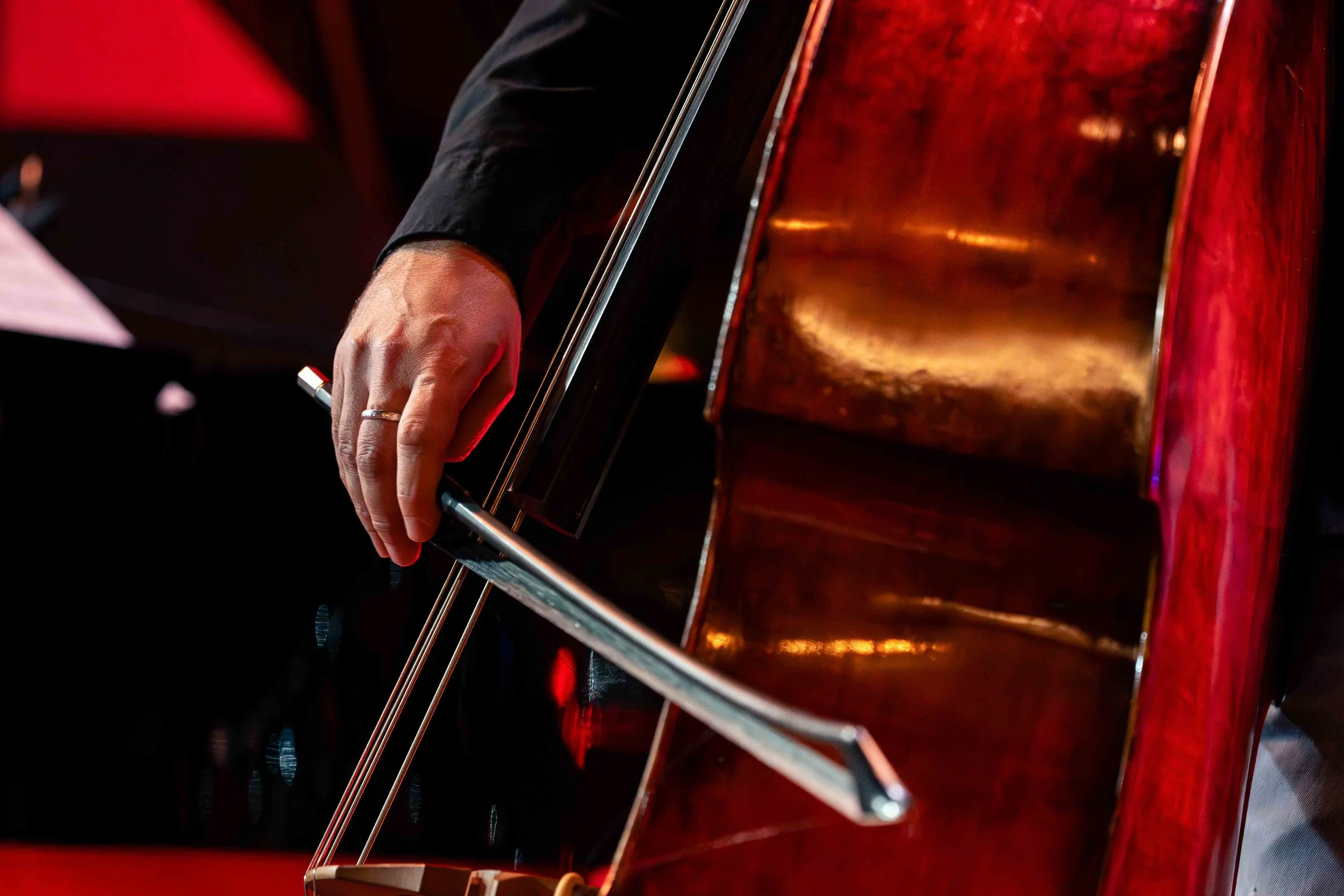 Photographie de reportage montrant un gros plan sur la main d’un contrebassiste jouant à l’archet lors d’un concert de jazz. Le cadrage met en valeur la texture du bois de l’instrument, le geste du musicien et la lumière rouge de scène qui baigne l’a