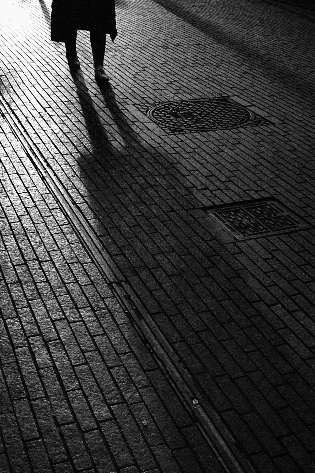 Photographie urbaine en noir et blanc montrant l'ombre allongée d'un passant sur un pavage éclairé par une lumière rasante à Bruxelles.