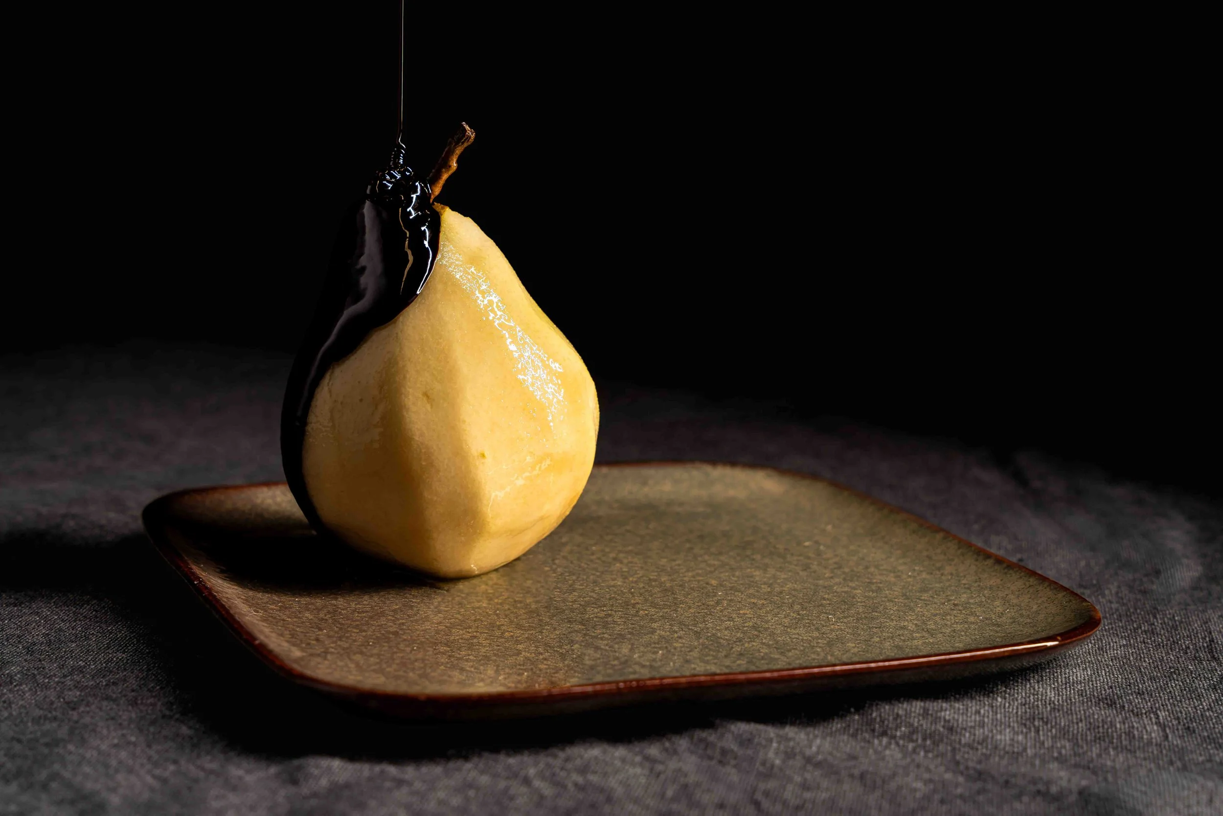 Photographie culinaire minimaliste montrant une poire pochée placée sur une assiette sombre, nappée d’un filet de chocolat coulant depuis le dessus. La lumière directionnelle met en valeur la texture du fruit et la brillance du chocolat dans une comp