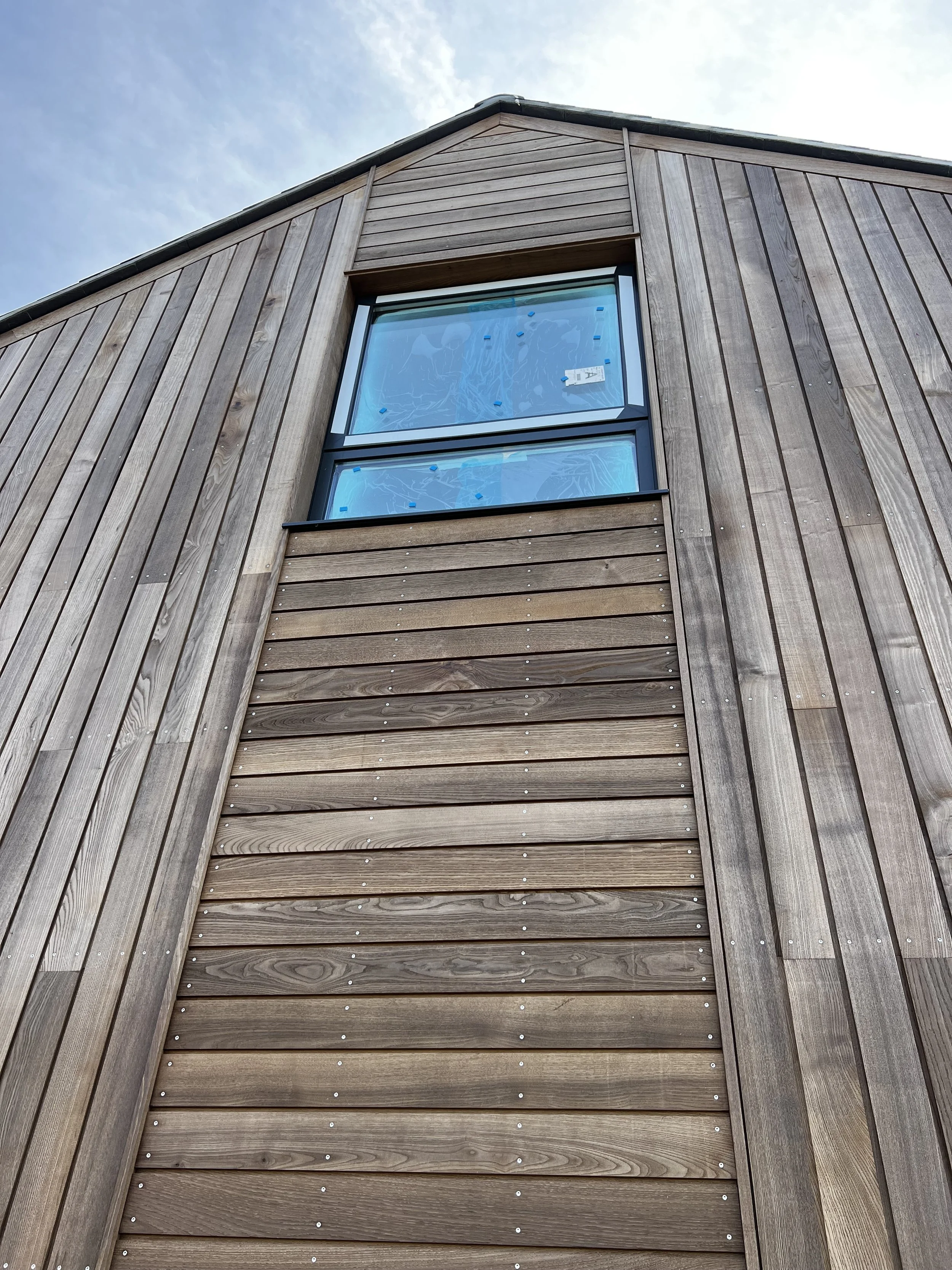 Close-up of a wooden building exterior with vertical and horizontal wooden panels and a large window on the upper floor, under a cloudy sky.