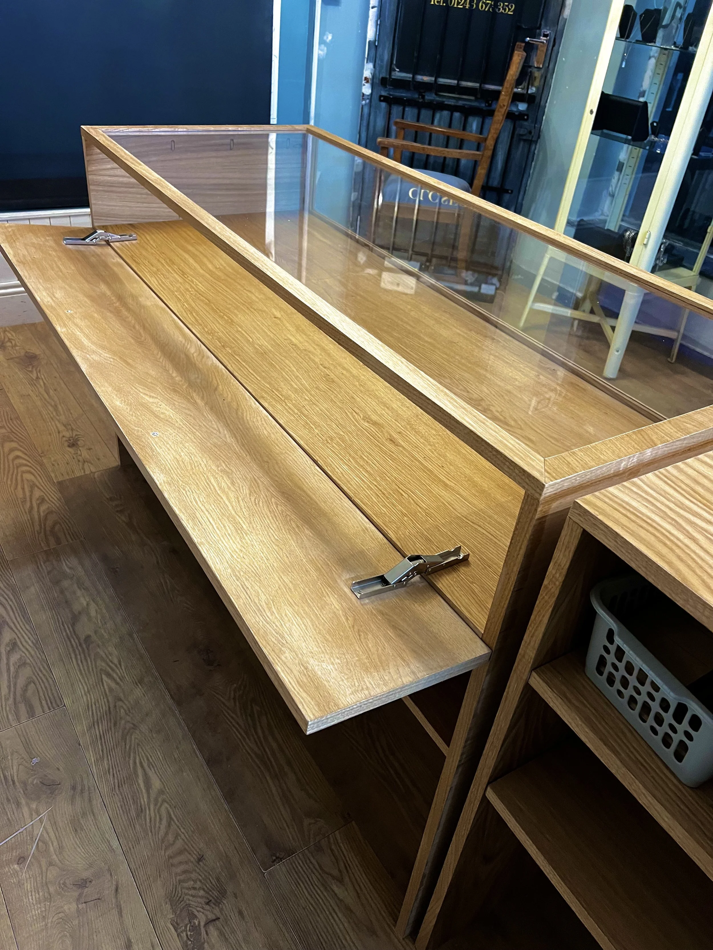 A wooden display case with a glass top on a wooden table, secured with metal fasteners, positioned in a retail or display store with wooden flooring and shelves in the background.