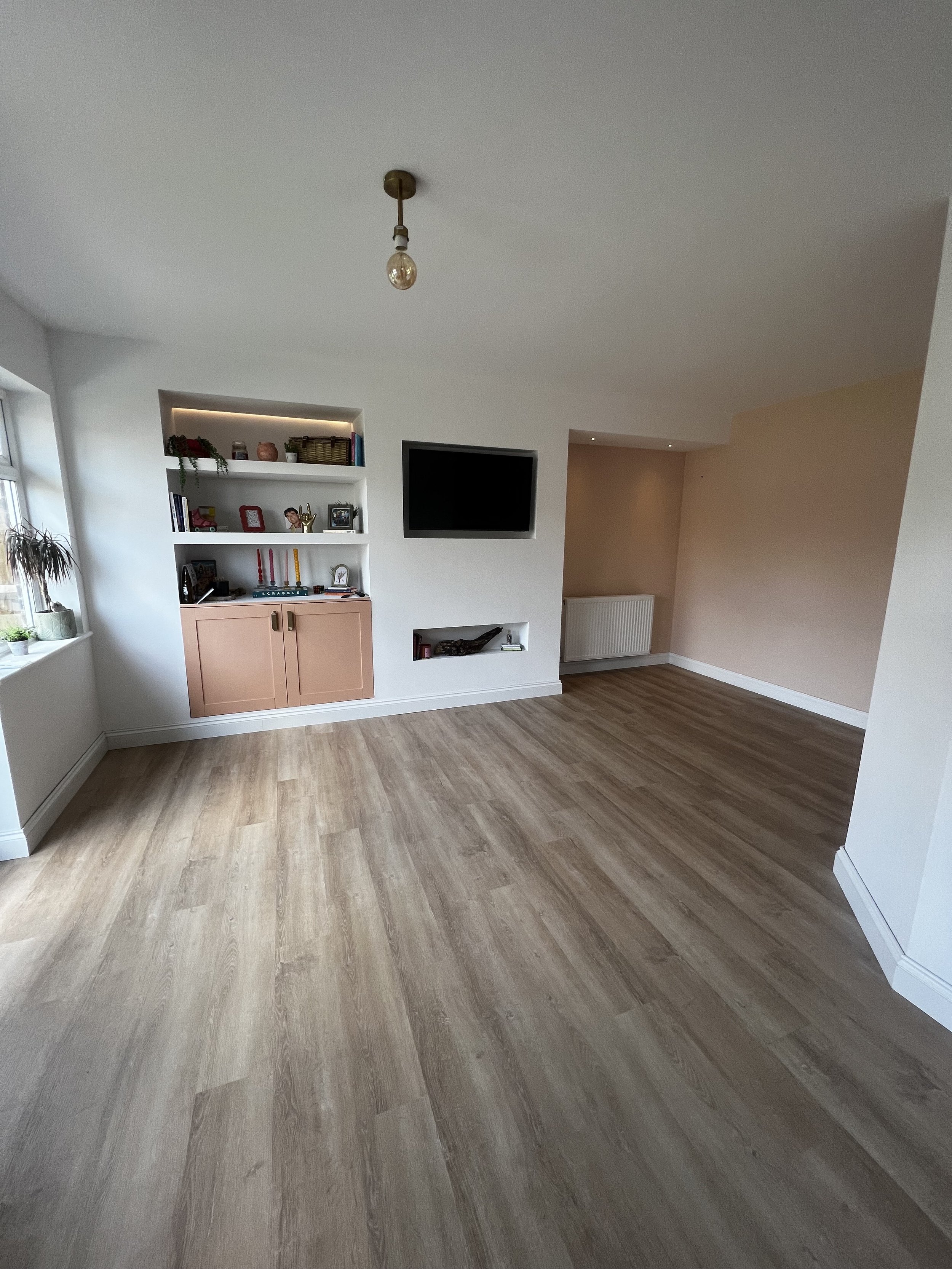 Empty living room with wood flooring, white walls, a built-in white shelf with pink doors, a wall-mounted TV, a window with plants on the sill, and a beige accent wall.