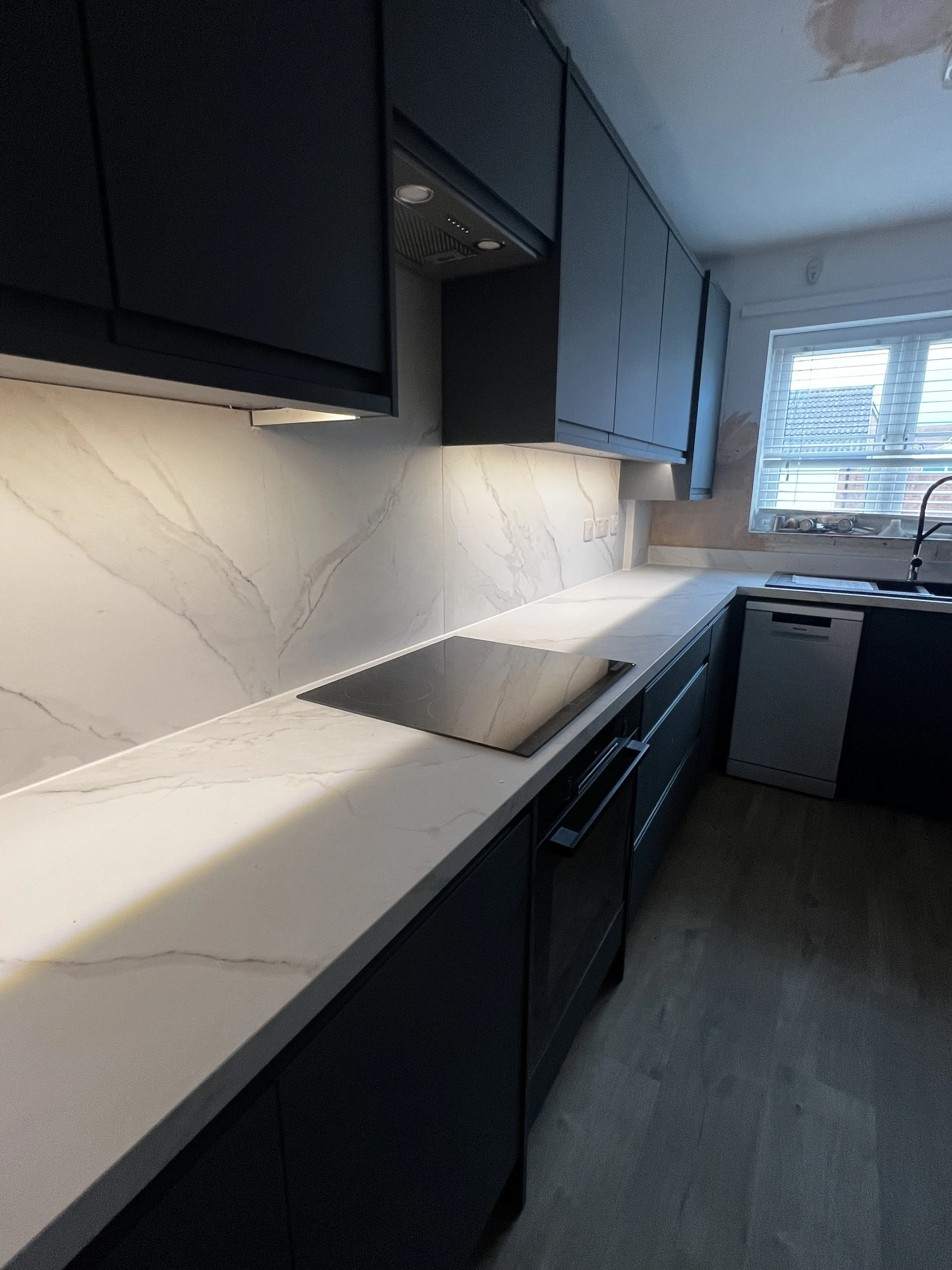 Modern kitchen with dark cabinets, white marble countertops and backsplash, a window with blinds, and a built-in stovetop.