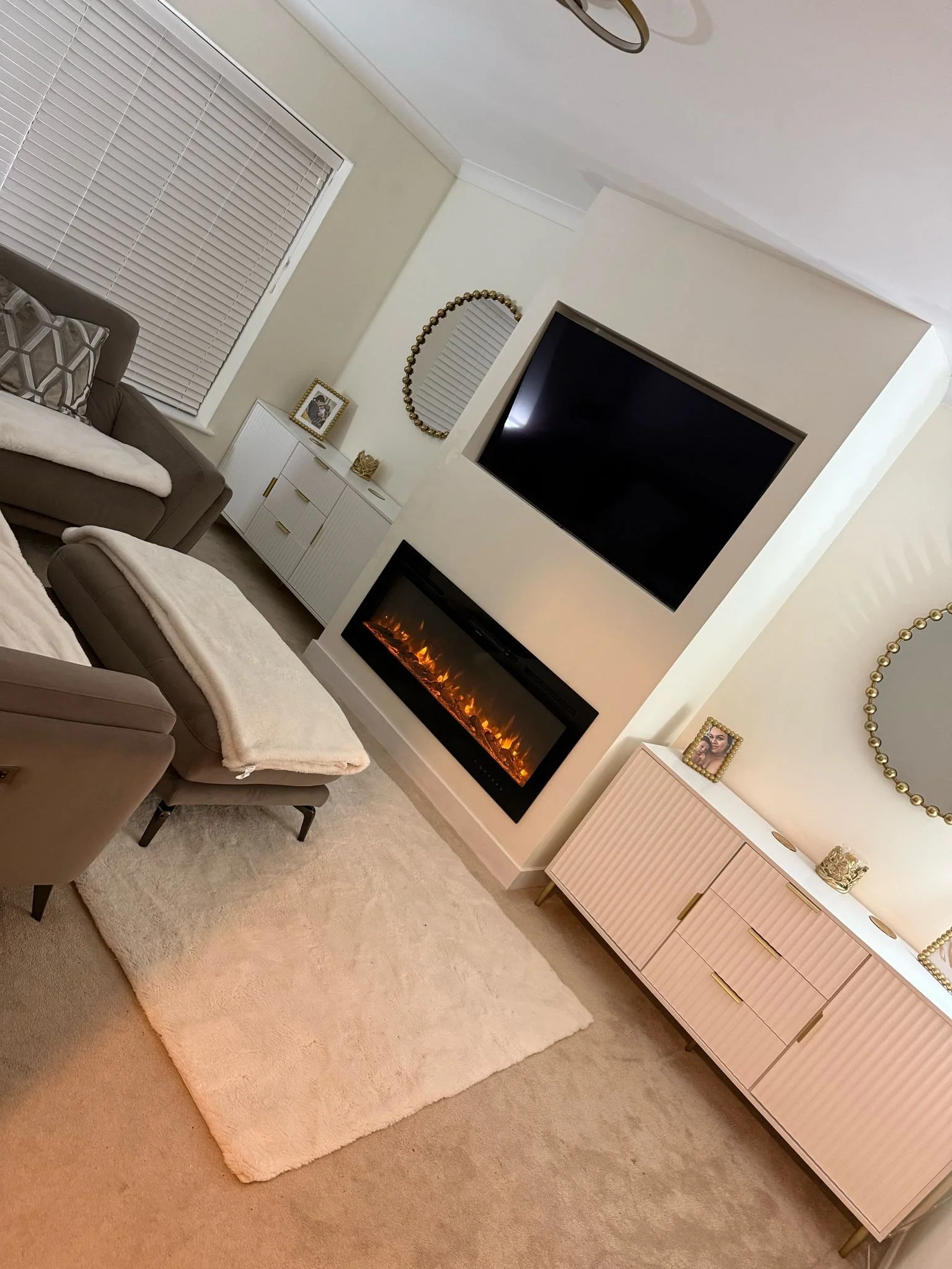 Living room with beige carpet, white shag rug, gray sofa with throw blankets and pillows, gray recliner with throw blanket, white sideboard with framed photo, gold decorative items, large wall mirror, flat-screen TV, electric fireplace, window with white blinds, ceiling light fixture.