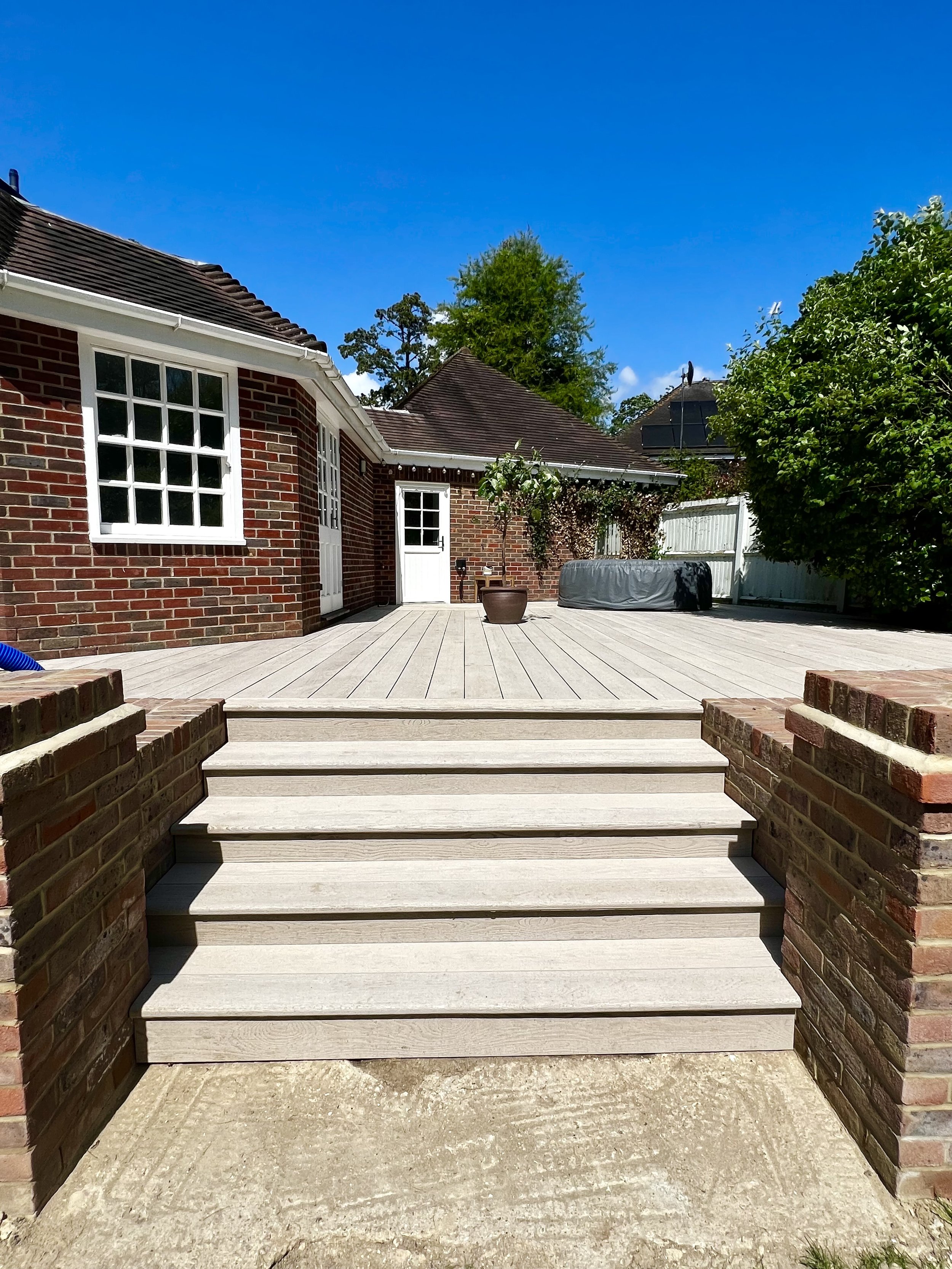 A backyard patio area with a light-colored wooden deck, stairs, and brick walls. There is a potted plant, a covered hot tub, and green trees with a clear blue sky overhead.