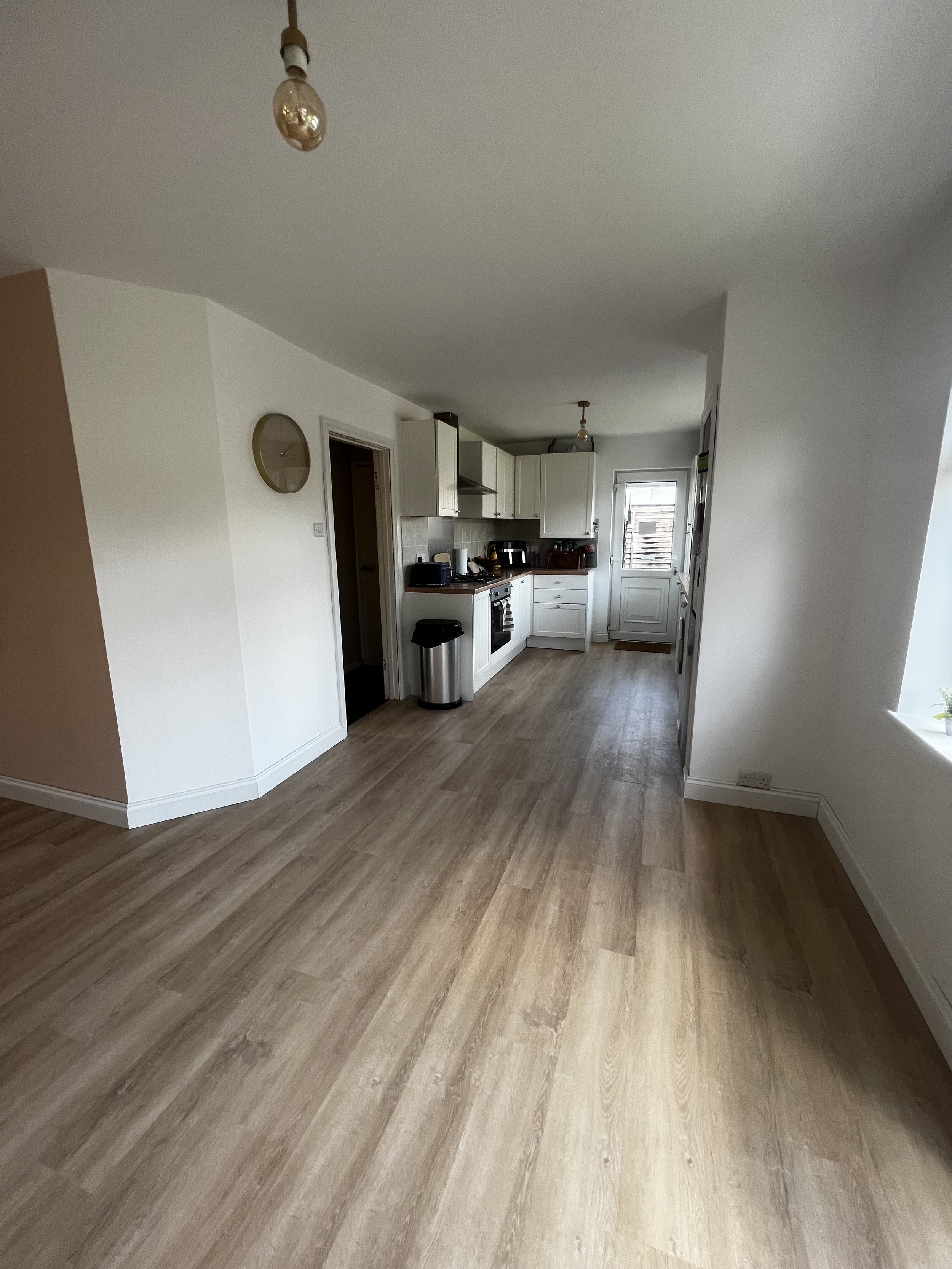 Empty kitchen and living space with light wood flooring, white cabinets, and a door leading to the outside. There is a wall clock and a light bulb hanging from the ceiling.