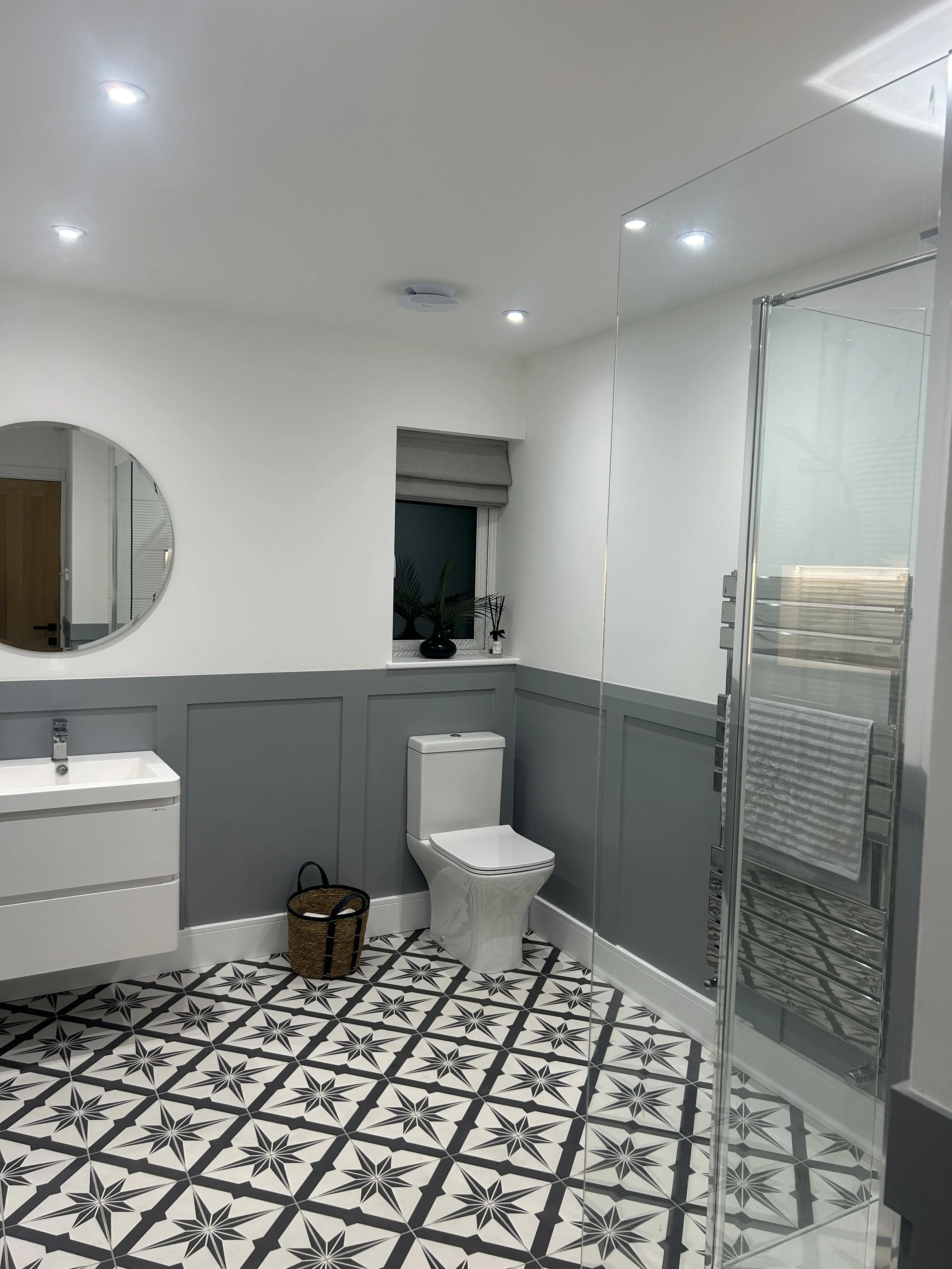 Modern bathroom with patterned black and white tile floor, white toilet, floating white sink, gray wainscoting, small window with gray shade, black vase with plant on windowsill, round mirror on white wall, basket on floor, and glass-enclosed shower.