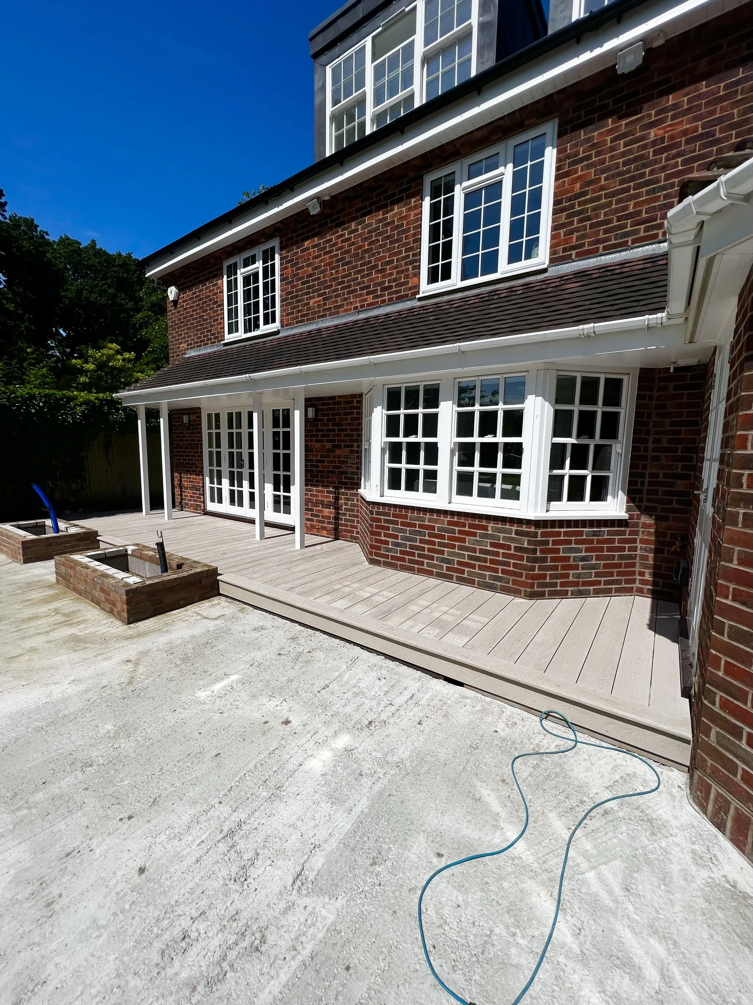 Backyard patio with a newly installed raised wooden deck adjacent to a brick house with multiple windows and a clear blue sky.