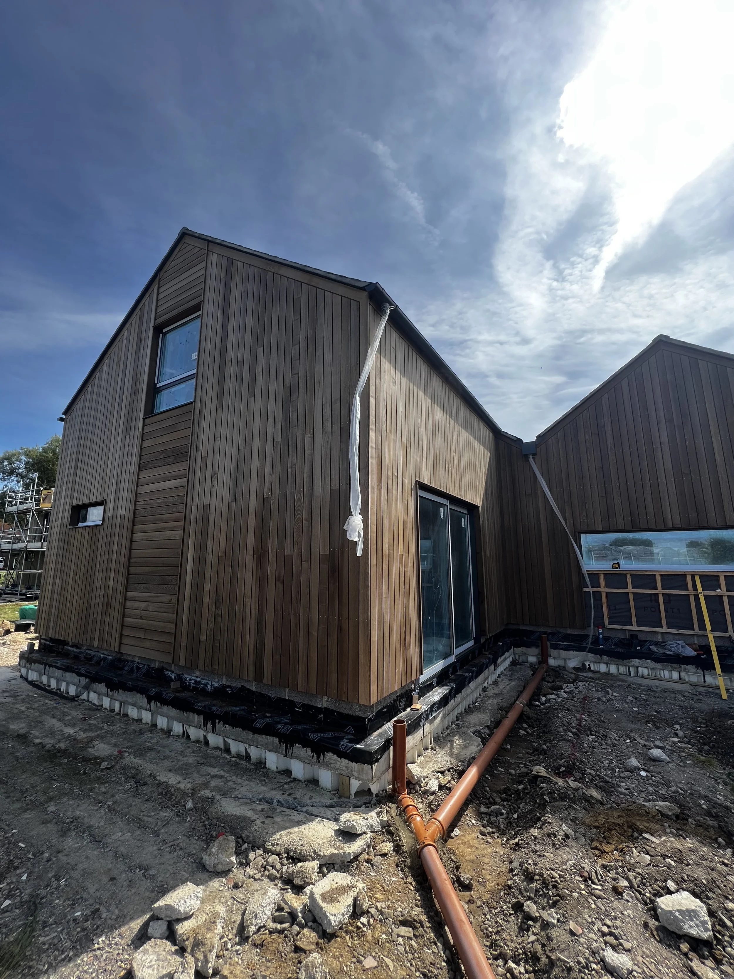 Newly built wooden house with construction materials and pipes visible at the base, under a partly cloudy sky.