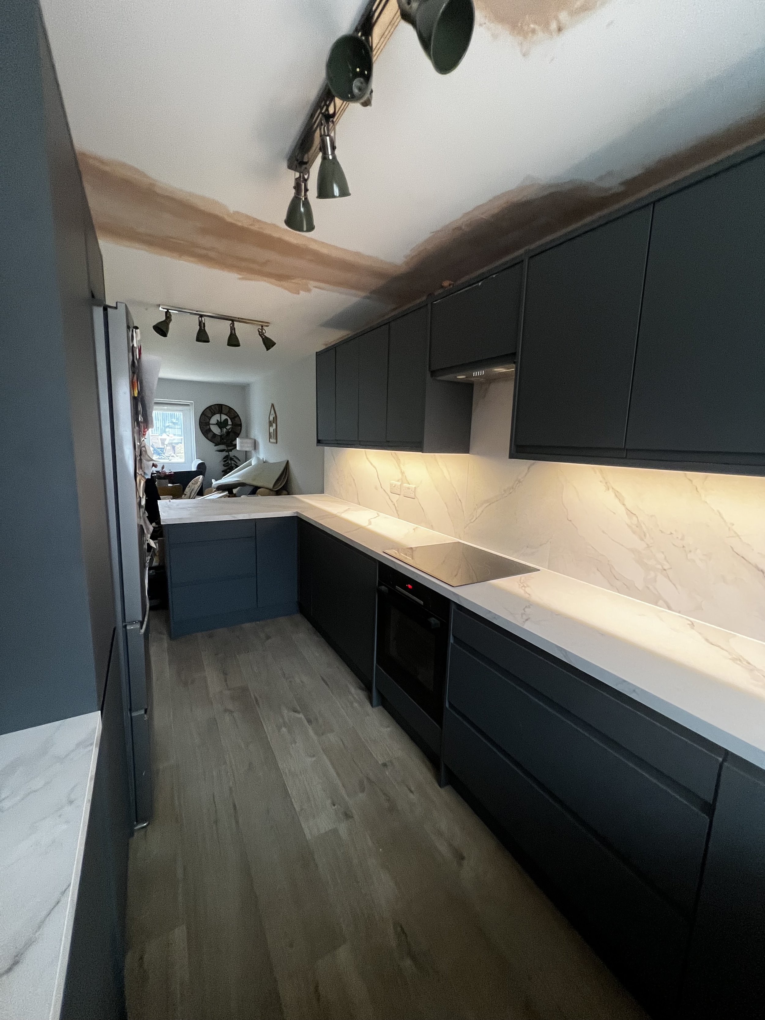 Modern kitchen with dark cabinets, marble countertops, and a marble backsplash under construction, with a ceiling light fixture and a view of the living room in the background.