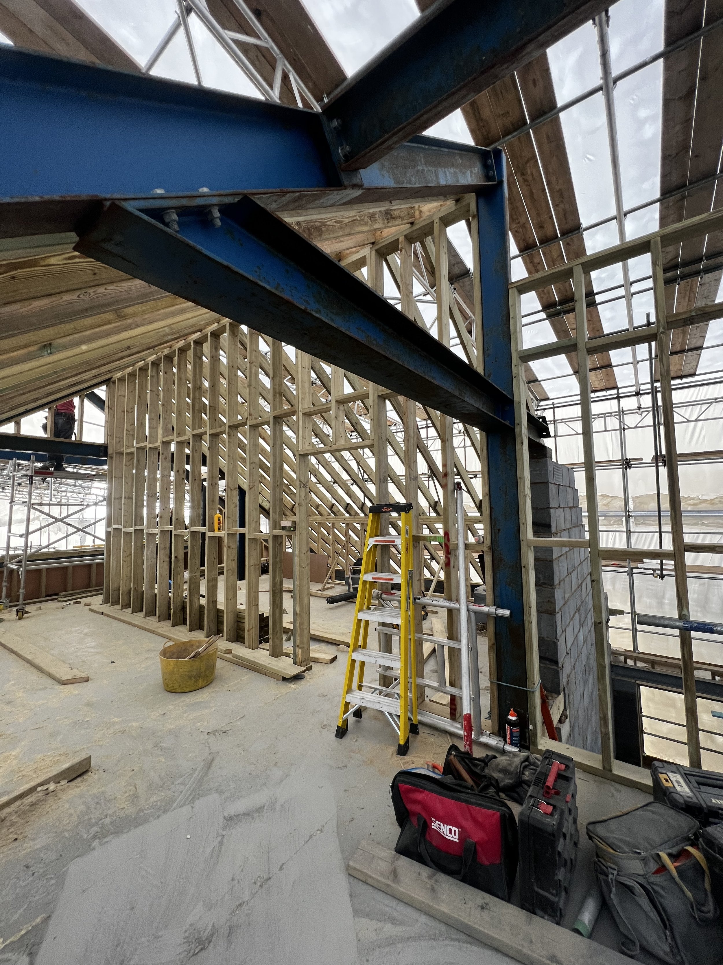 Construction site with wooden framing, a yellow ladder, tools, and a metal beam structure in an unfinished building.