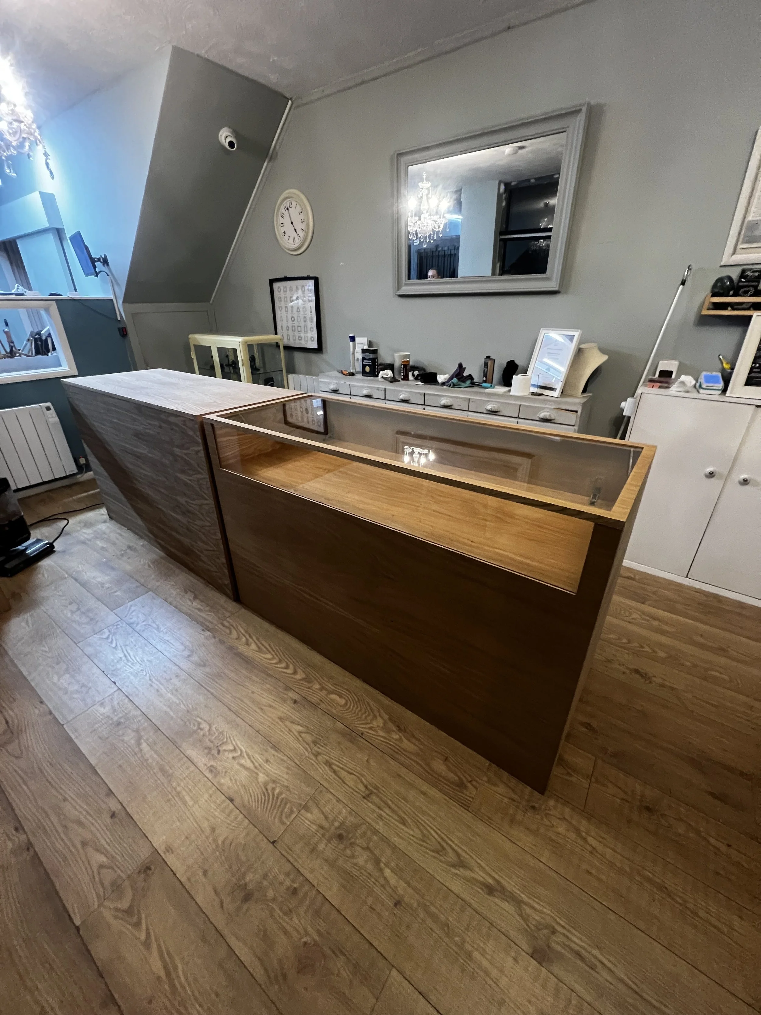 A wooden display counter with a glass top in a room with light gray walls, a large mirror, and various items on a white dresser behind it.