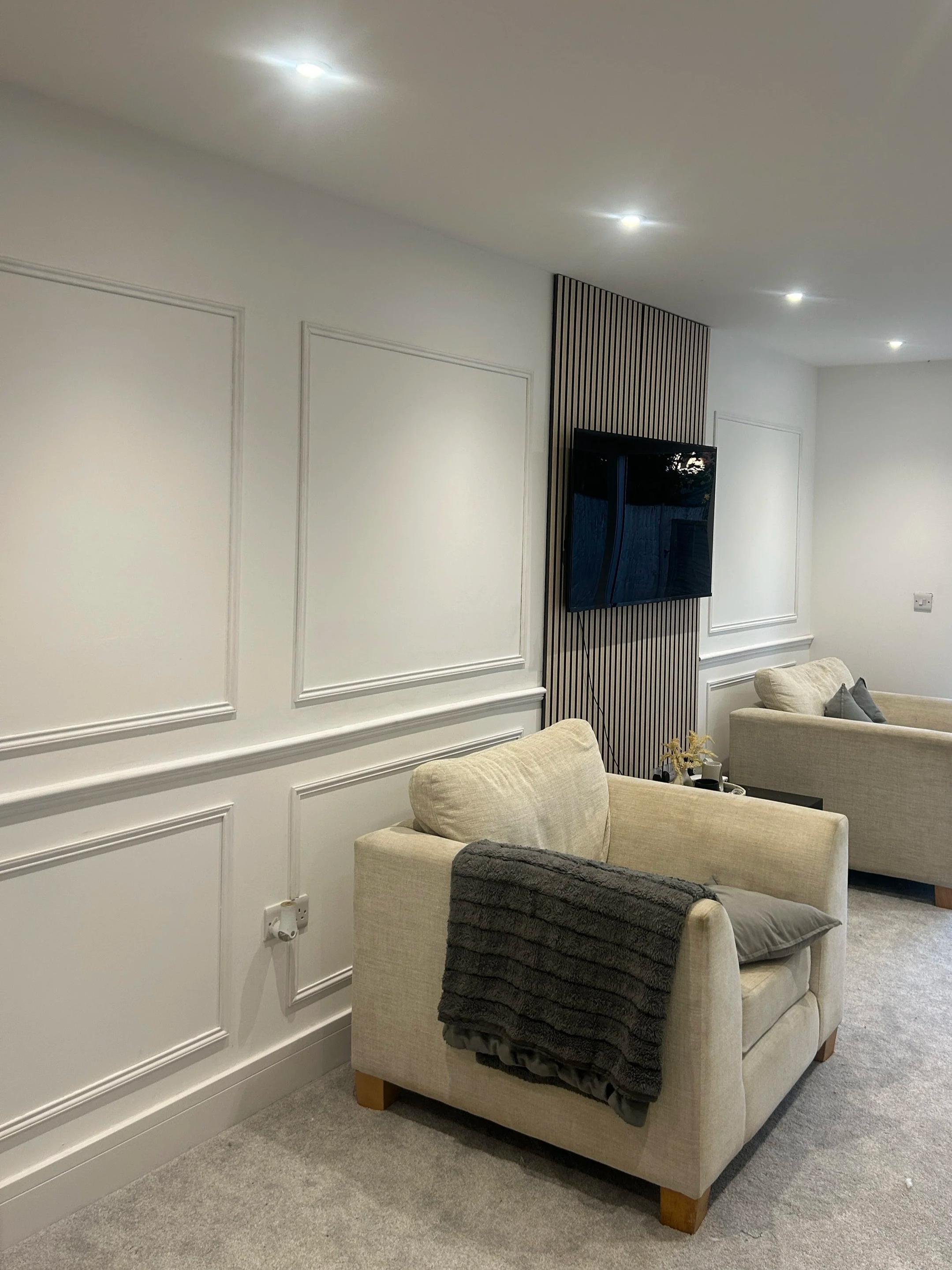 Living room with beige armchair, throw blanket, and gray pillow, two beige sofas, wall paneling, mounted flat-screen TV, and carpeted floor.