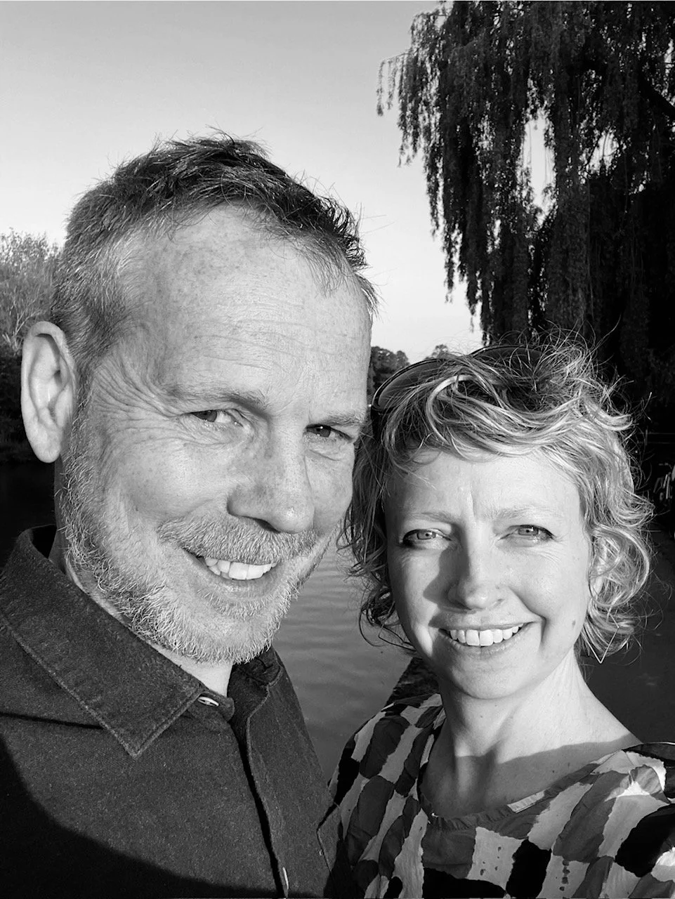 A black and white photo of a smiling man with short hair and beard and a woman with curly short hair, outdoors near a body of water, with trees in the background.