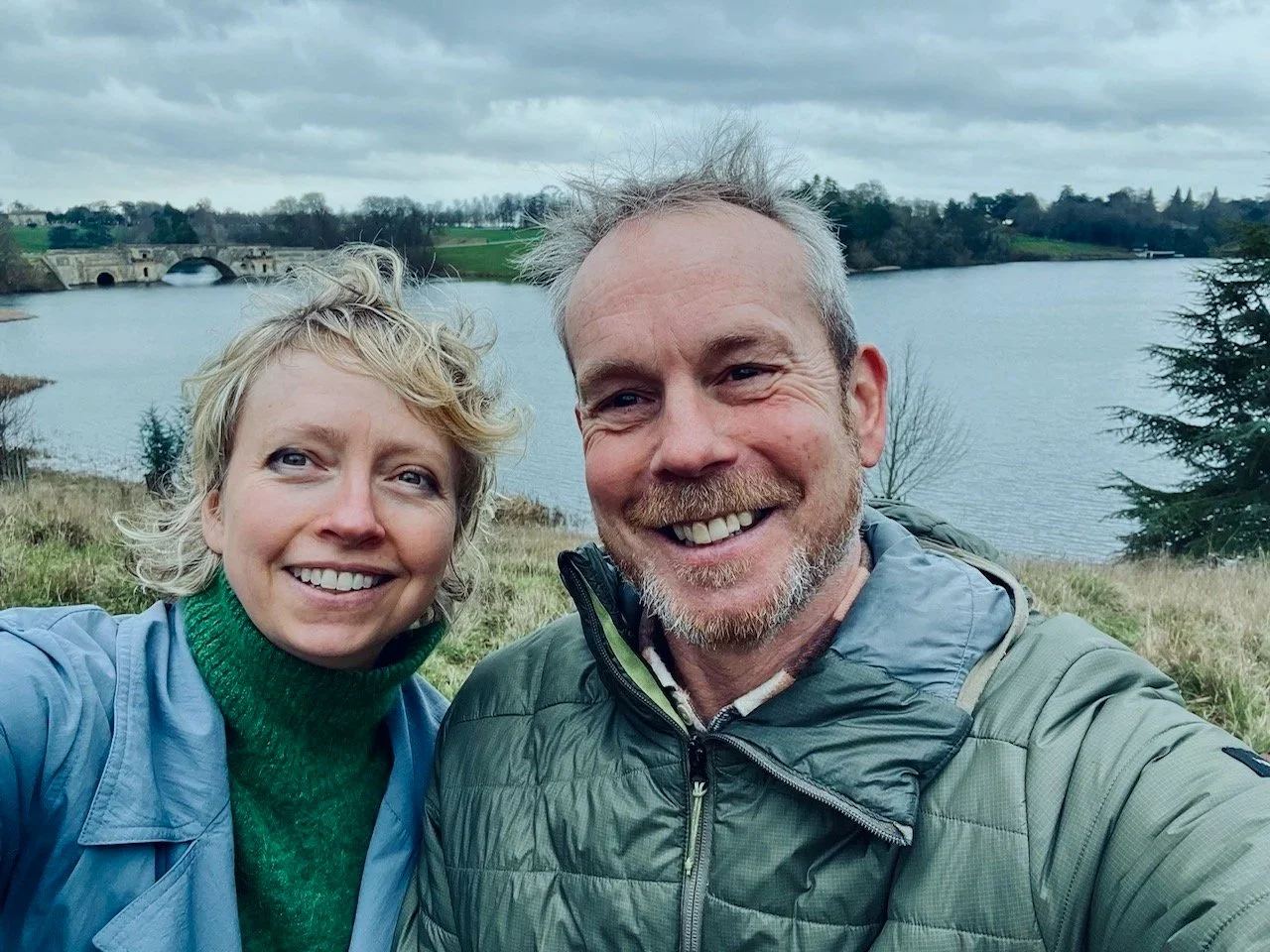 A smiling man and woman taking a selfie outdoors near a river with a bridge and trees in the background, under a cloudy sky.