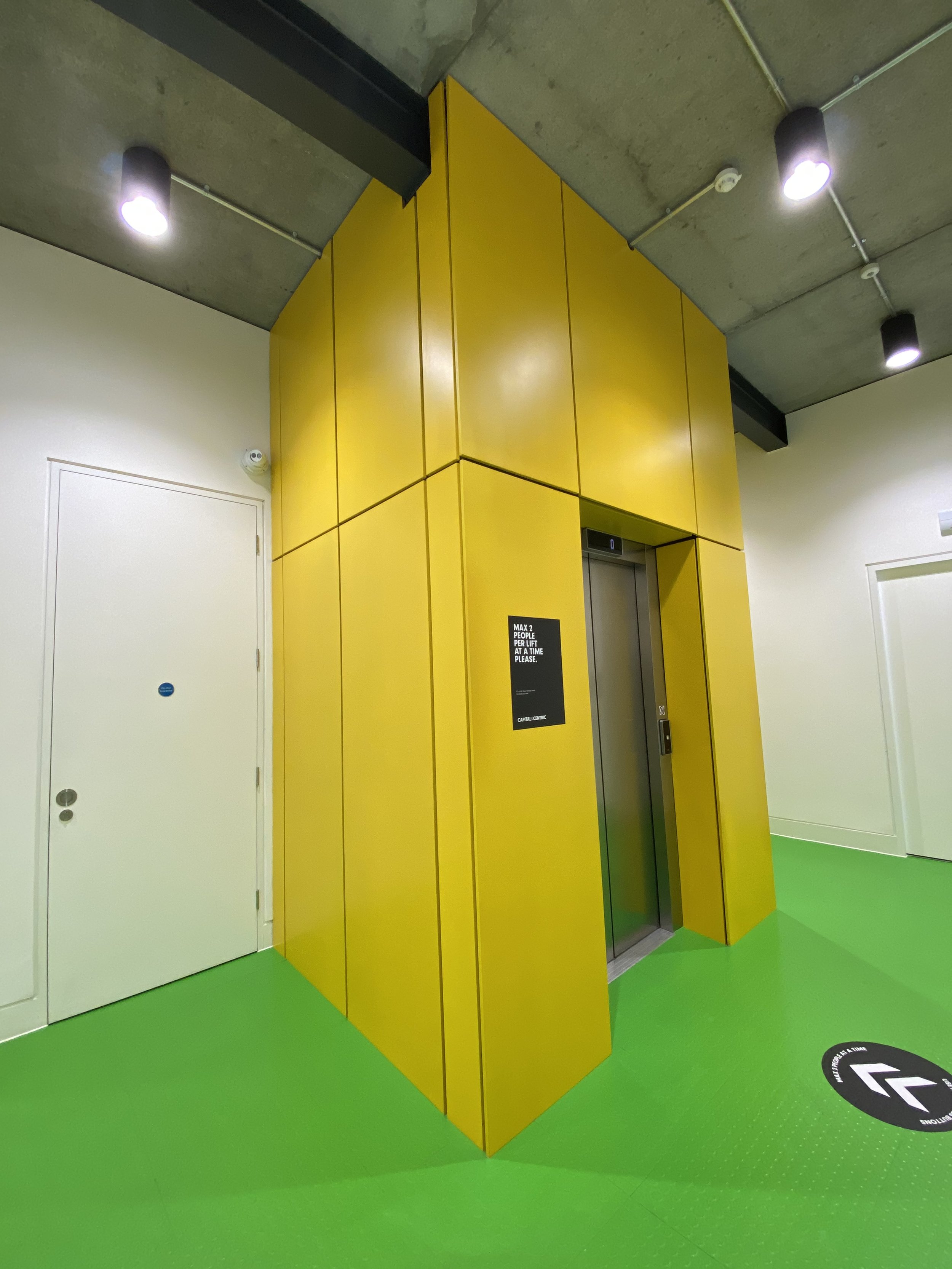 Elevator with yellow panels and a black sign, set in a green floor area with directional markings, in a modern indoor space.