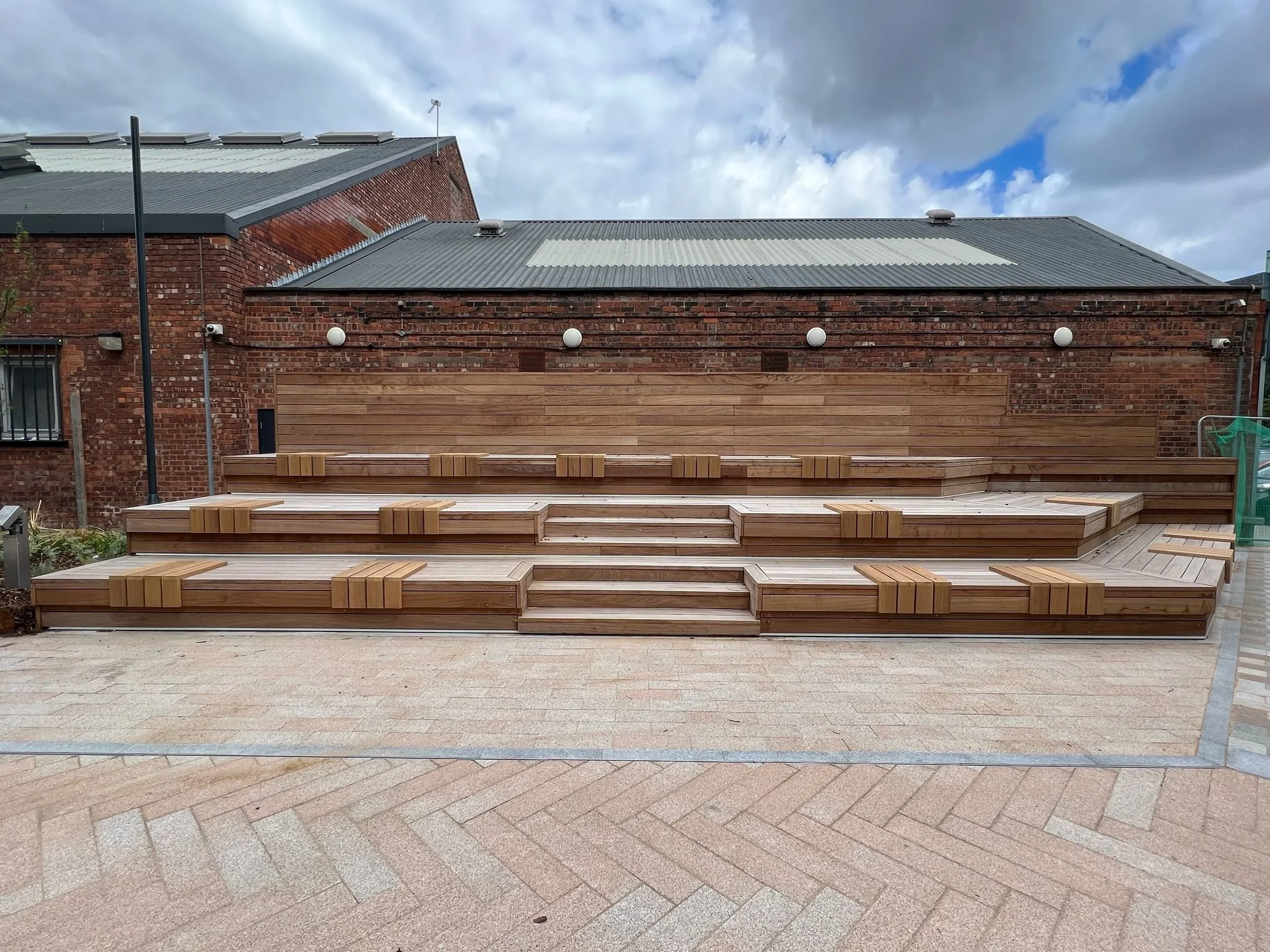 Wooden tiered seating structure in an outdoor area with brick buildings and a cloudy sky in the background.