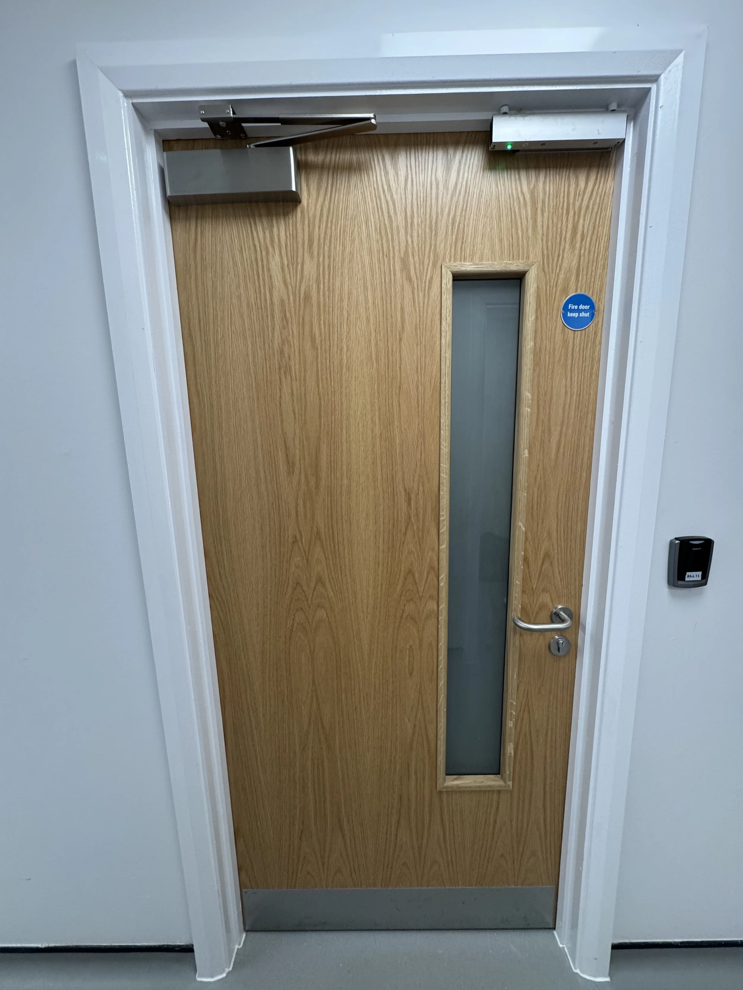 Wooden fire door with a narrow vertical window, a metal push bar handle, and a blue sticker that says 'Fire door keep shut,' surrounded by white trim and a gray wall.