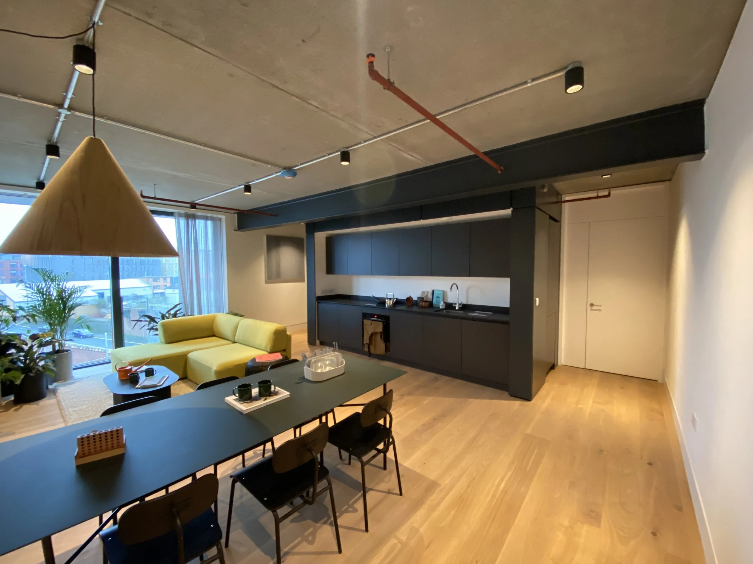 Open-plan living room and kitchen with yellow sectional sofa, black kitchen cabinets, wooden dining table with chairs, large window with city view, indoor plants, and minimalist decor.