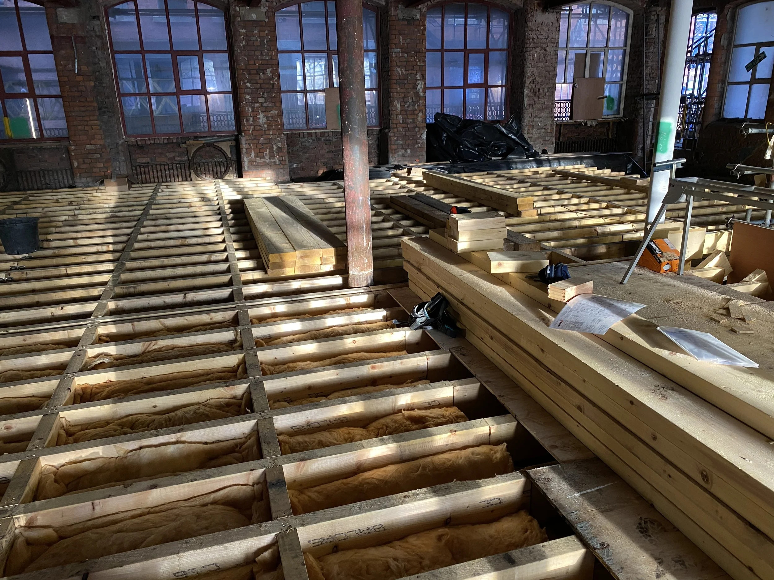 Construction site indoors with wooden floor framing and insulation, construction tools and materials in an industrial building with large windows and brick walls.
