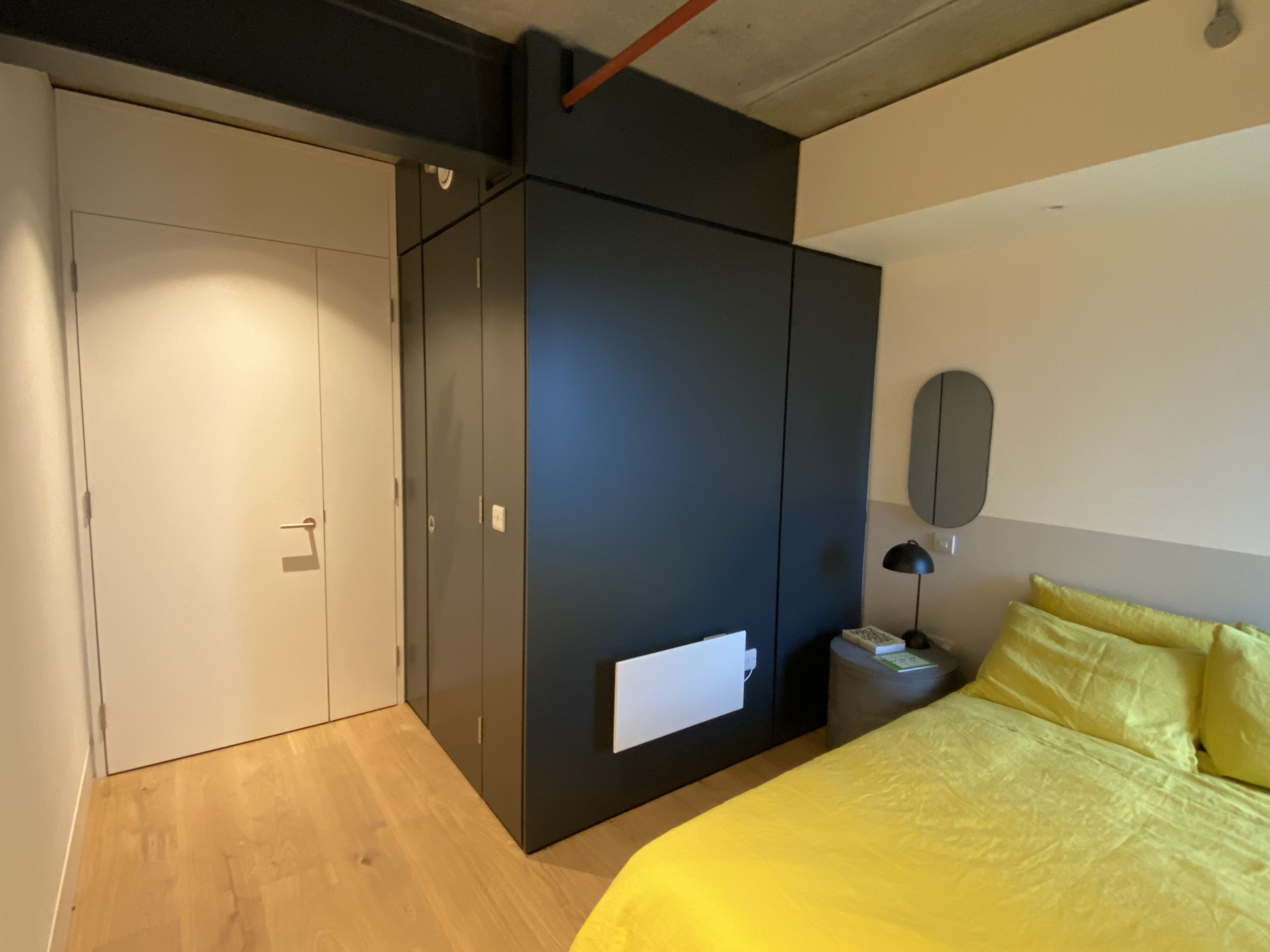 A bedroom with a yellow bedspread and pillows, a bedside table with a lamp and books, and a black wardrobe. The walls are light-colored, and there is a mirror and a heater on the wall.