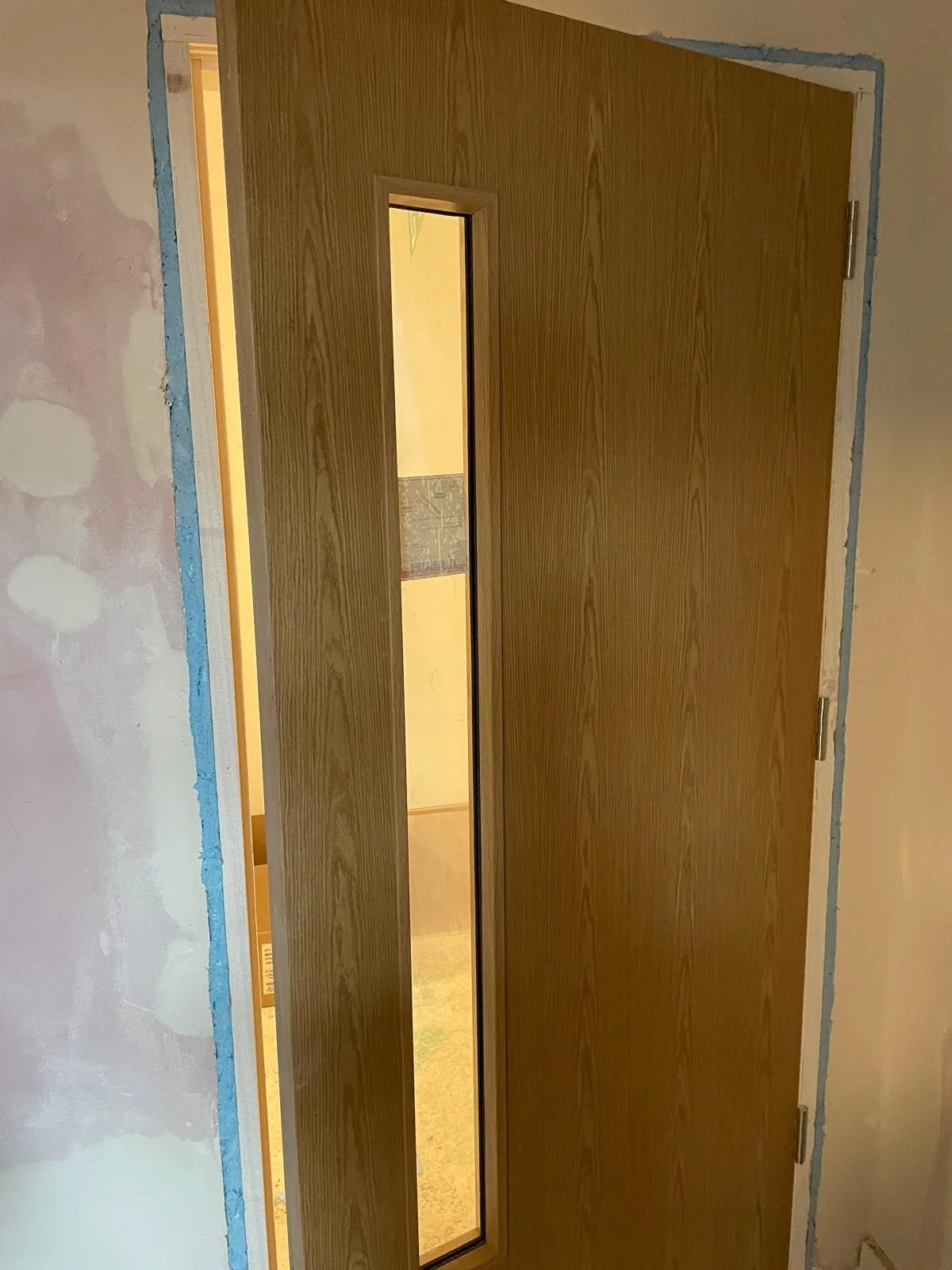 Wooden door with a vertical glass window, partially open, in a room under renovation.