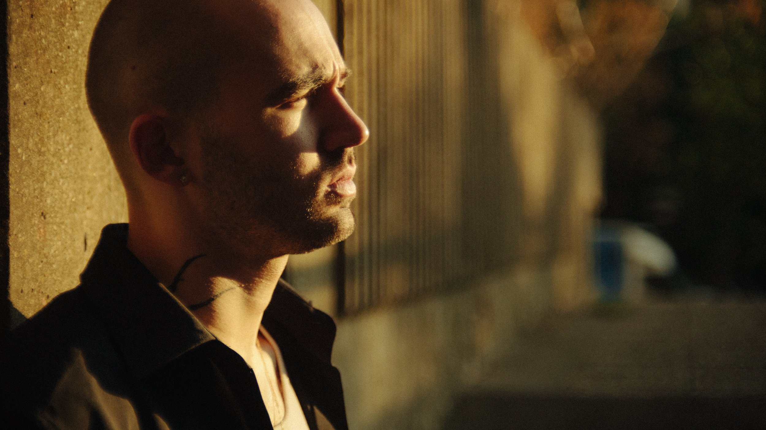 A young man with a shaved head and stubble, facing left with closed eyes, is standing against a wall during sunset or sunrise.