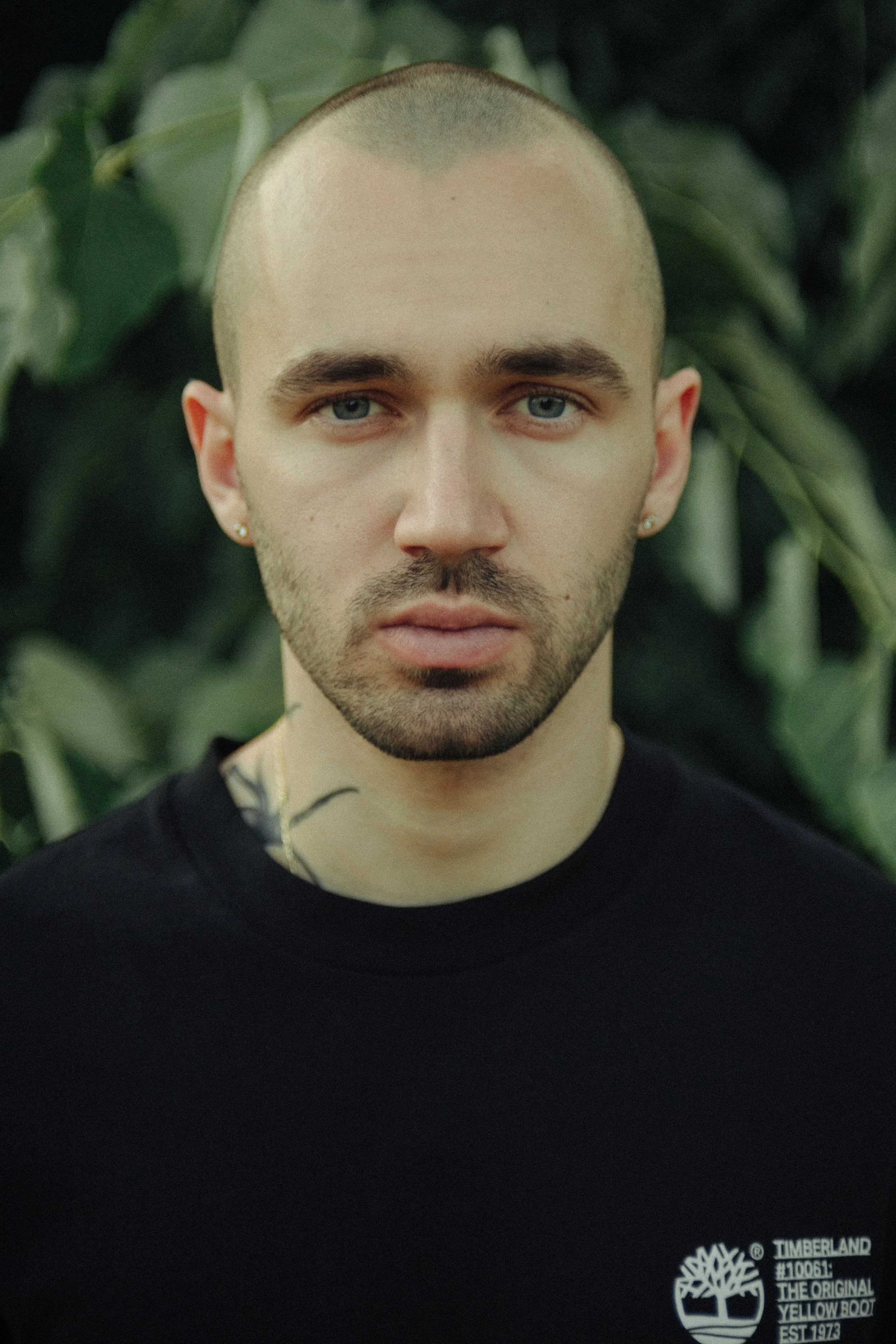 A young man with short hair and blue eyes wearing a black Timberland t-shirt, standing in front of green foliage.