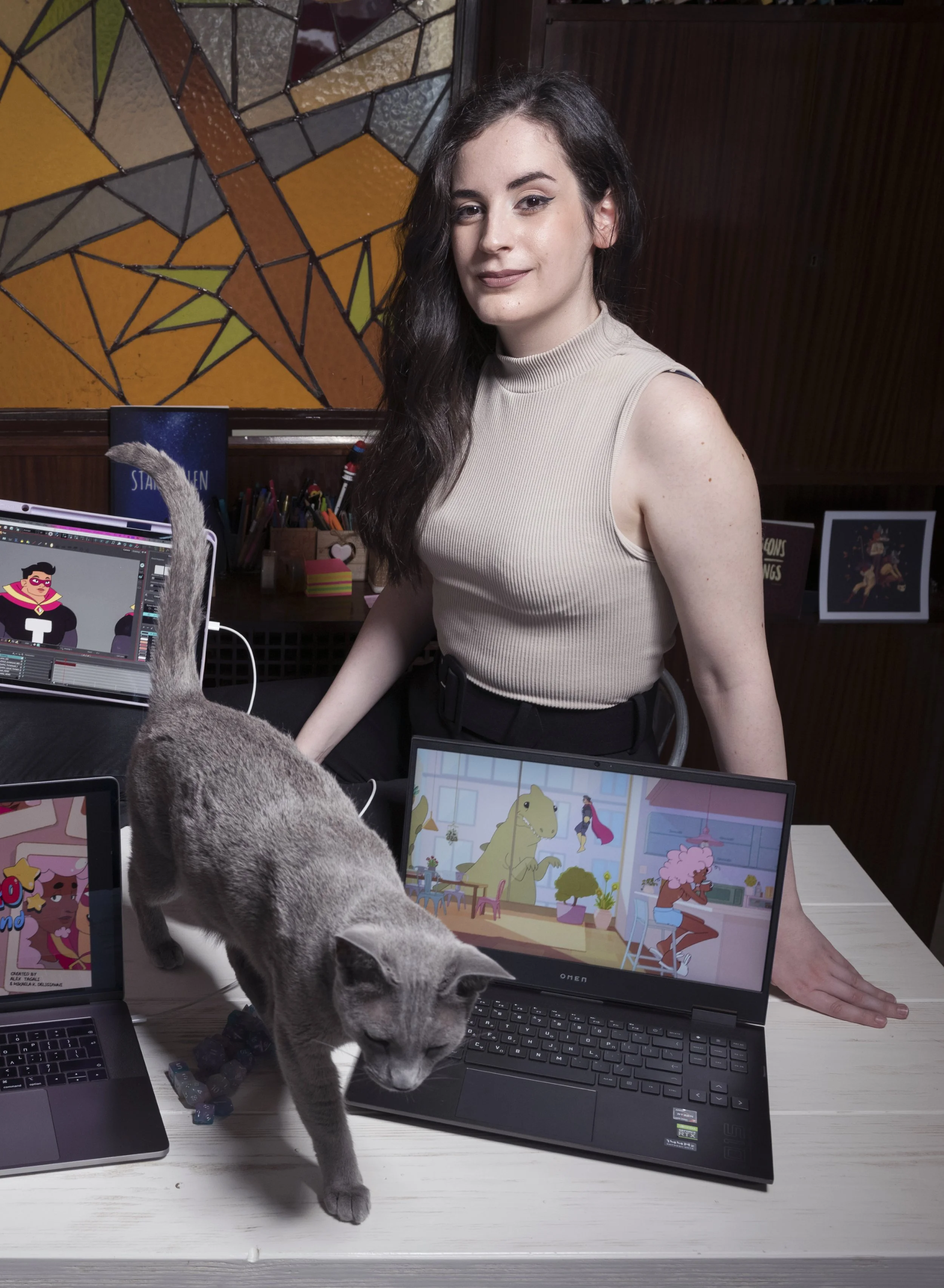 A young woman with long dark hair and light skin standing at a wooden table, surrounded by laptops displaying animated characters, a gray cat walking across the table, and a stained glass wall panel in the background.