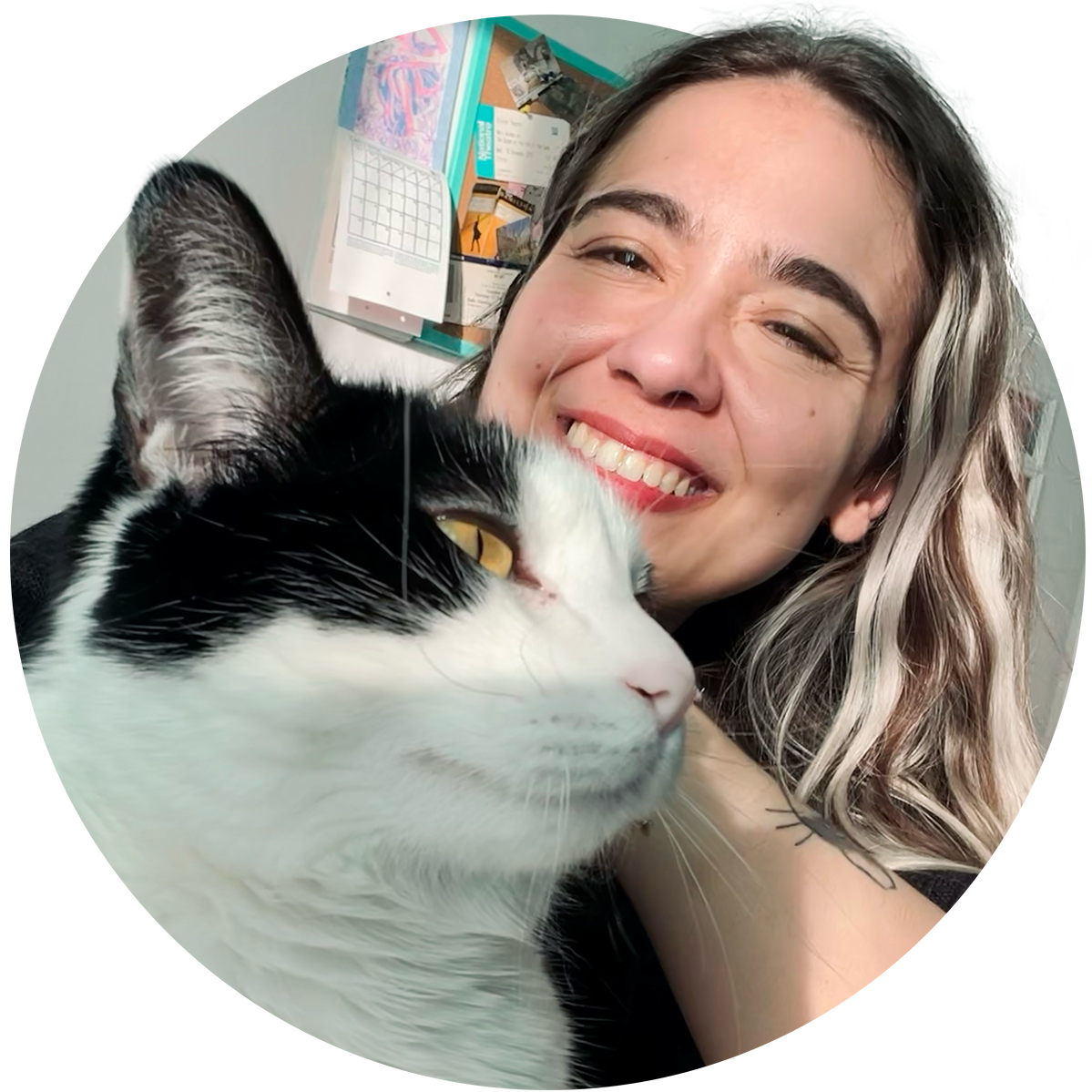 A young woman with wavy hair and a bright smile takes a selfie with a black and white cat in front of her, indoors with a bulletin board in the background.