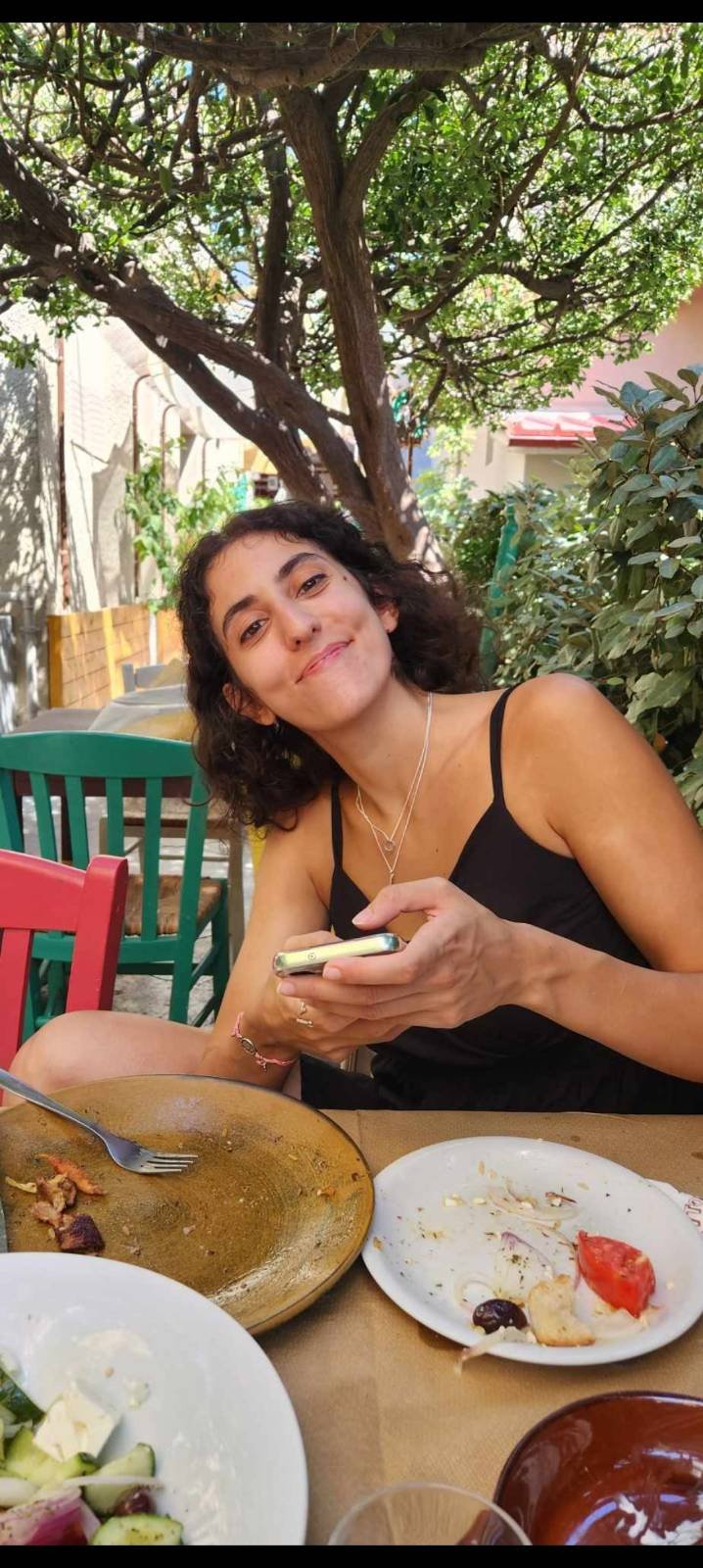 Young woman with curly dark hair in black tank top sitting outdoors at a table, holding a smartphone, smiling at the camera, with trees and garden in the background.