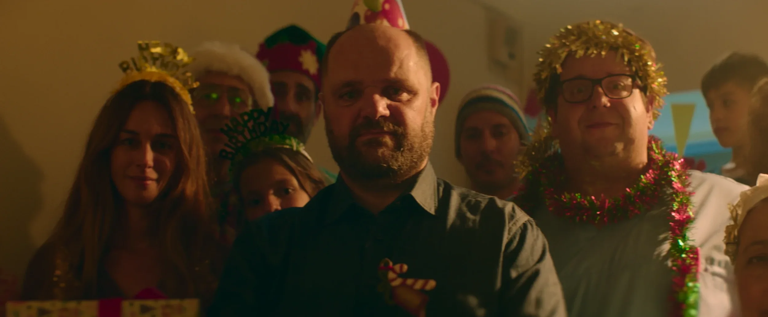 A group of people celebrating a birthday party, wearing party hats, tinsel, and festive decorations, with some smiling and looking at the camera.