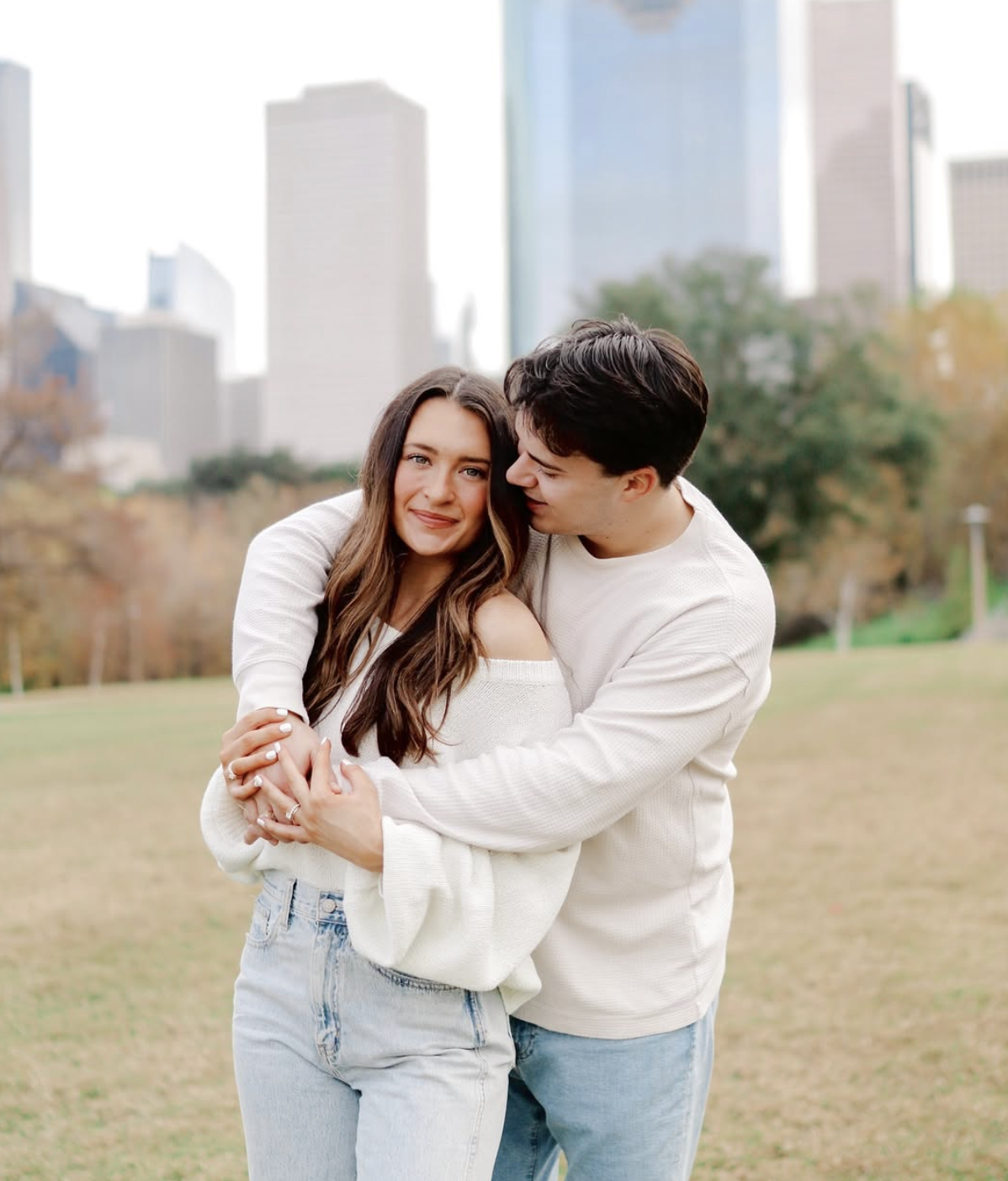 A couple hugging outdoors in a park with city skyscrapers in the background.