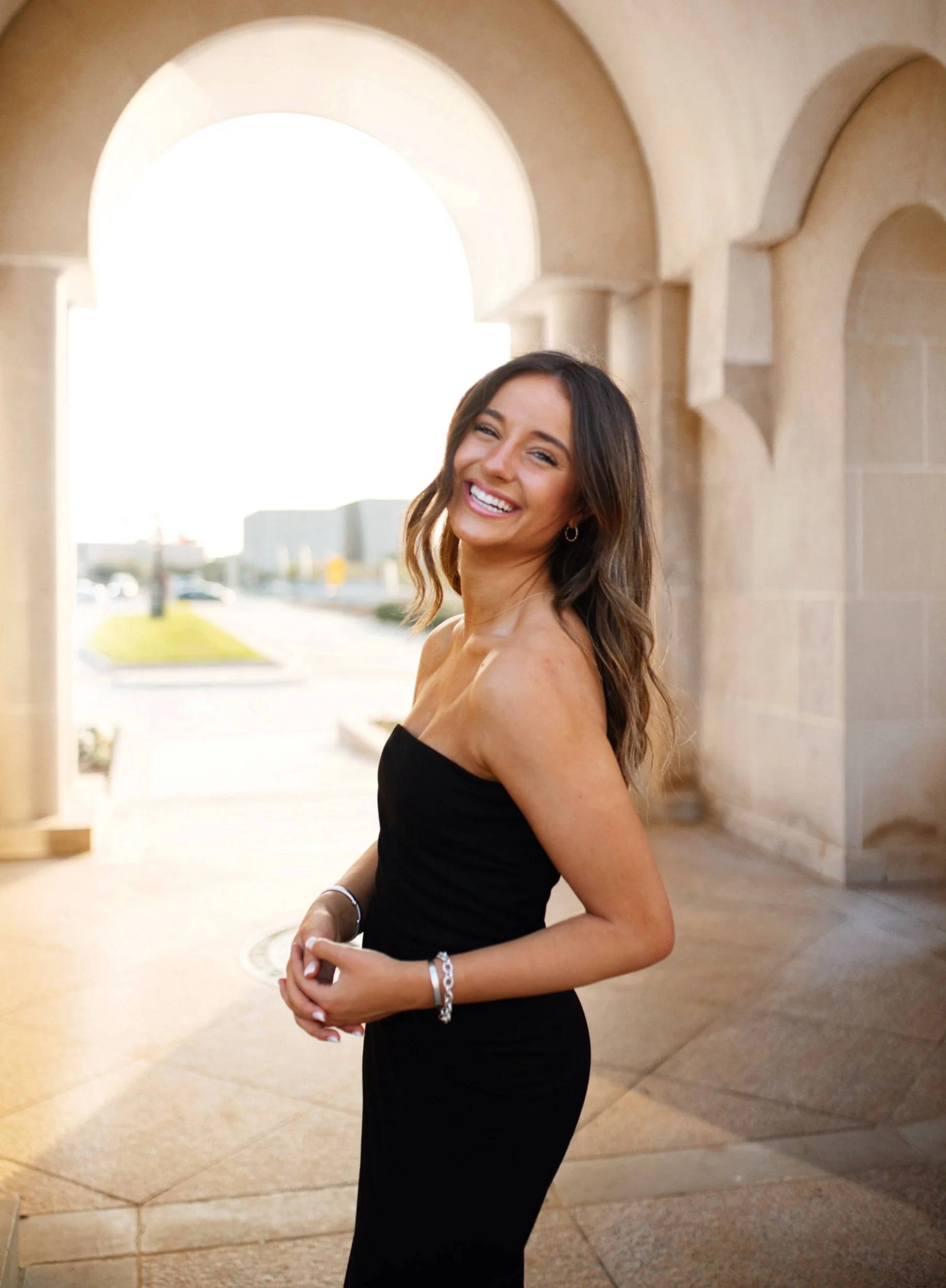 A smiling woman in a black strapless dress poses outdoors near a stone archway, with a sunny background and modern buildings in the distance.